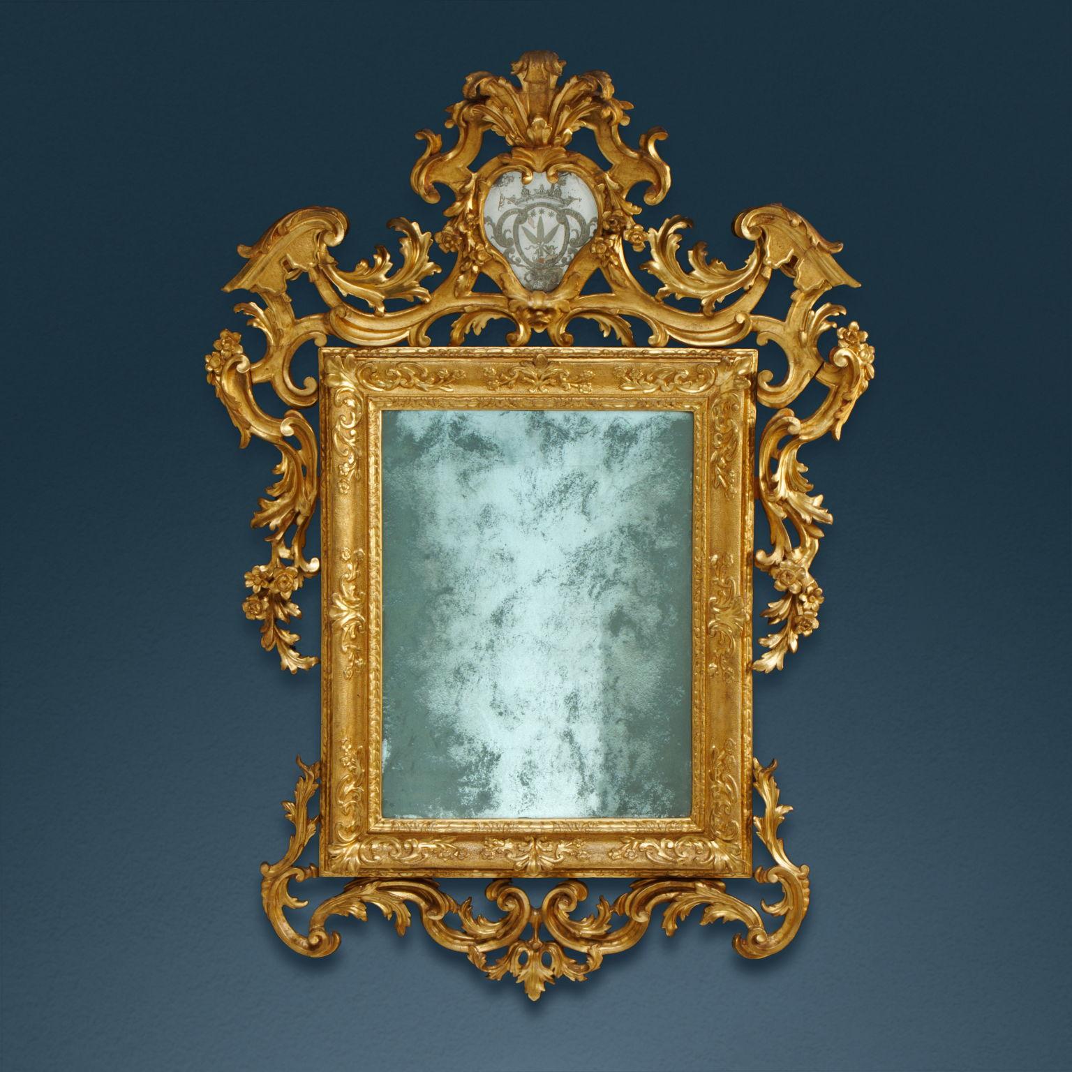 Carved and gilded wooden mirror. The rectangular frame enclosing the mirror is carved with small scroll motifs on the corners with the bottom tapped with a burin. Surrounding the frame is a carved decoration with architectural volutes and foliage