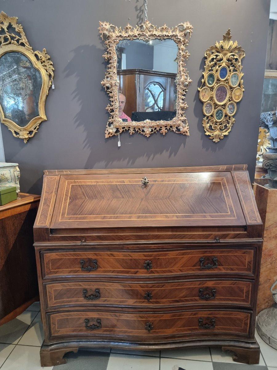 Specchiera veneziana della seconda metà dell’Ottocento, interamente intagliata a mano, laccata in policromia e dorata a guazzo. La cornice è un autentico capolavoro di maestria artigianale, con un decoro che simula un merletto traforato, dove le