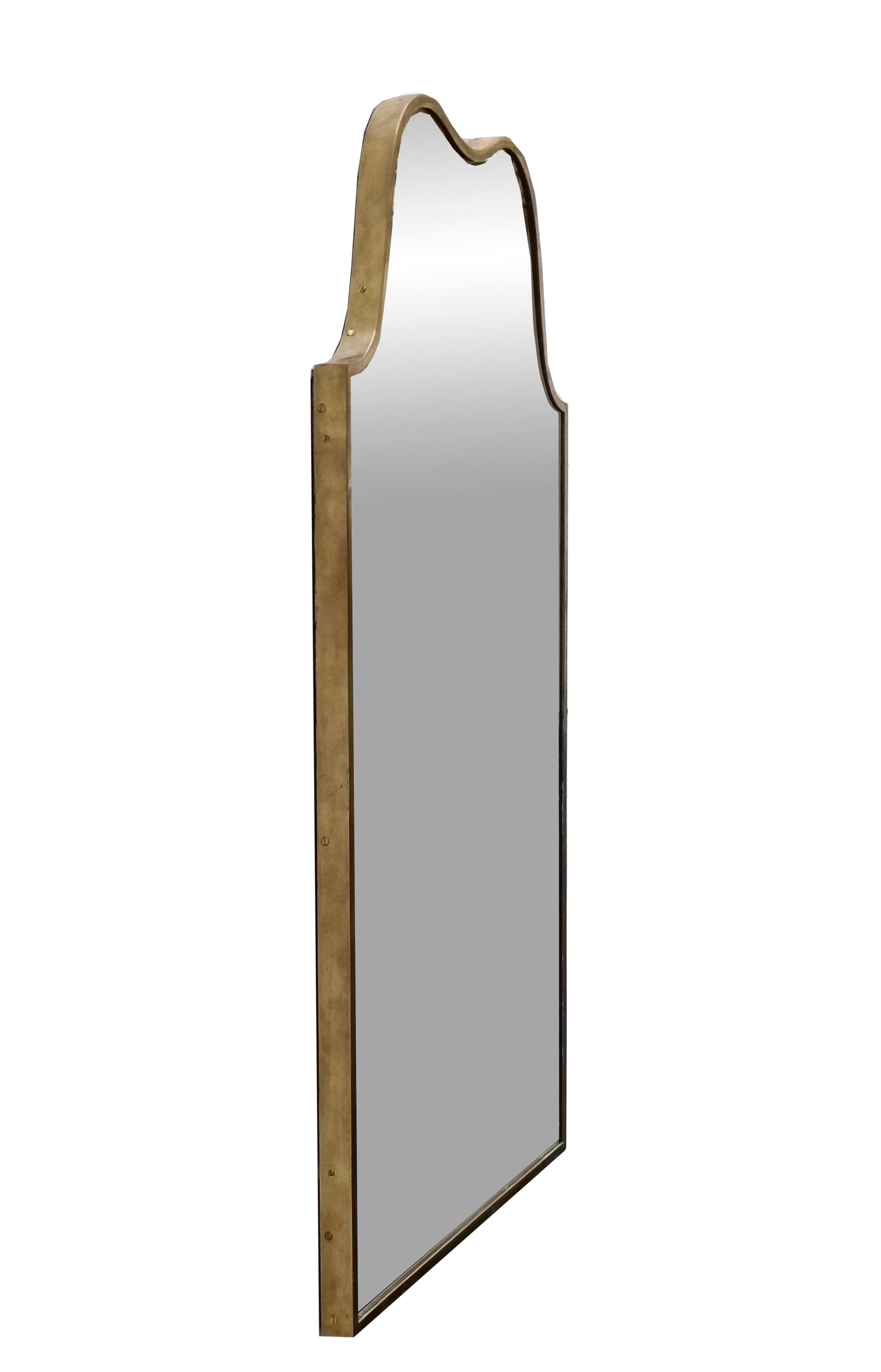 Este elegante espejo de pared italiano de mediados de siglo presenta una grácil silueta  enmarcado por un fino borde de latón que realza su refinada sencillez.Espejo de pared con una forma sinuosa y un diseño curvado en la parte superior que evoca