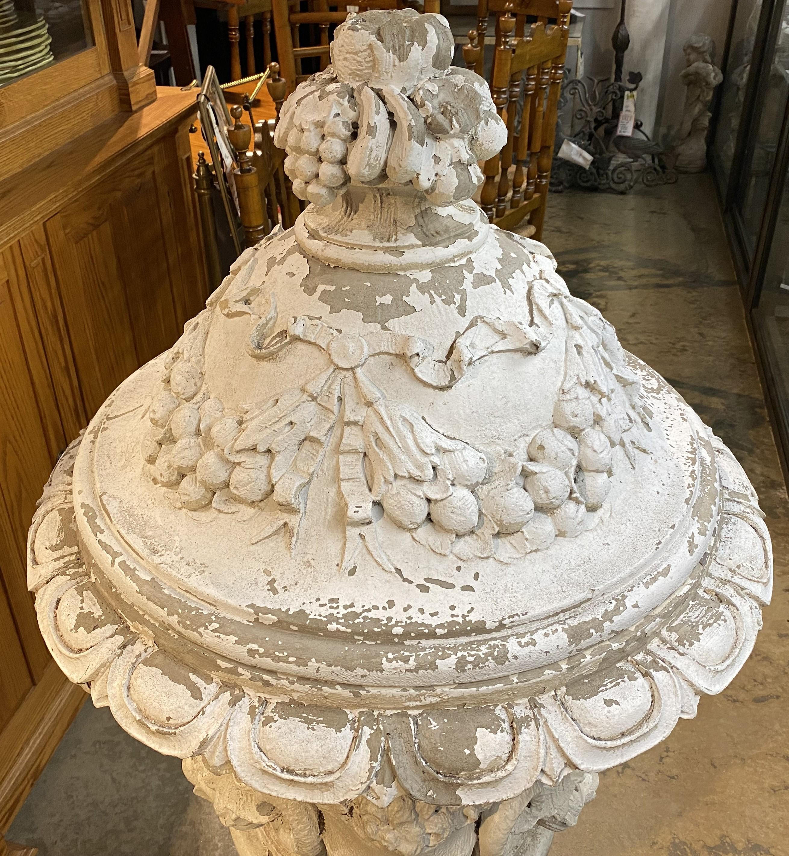 Spectaculaire urne de jardin couverte de forme classique en fausse pierre, avec décor de têtes de bélier, de fruits et de ruban. Fabuleuse patine d'ensemble avec des pertes de peinture et des imperfections correspondant à l'âge et à l'utilisation.