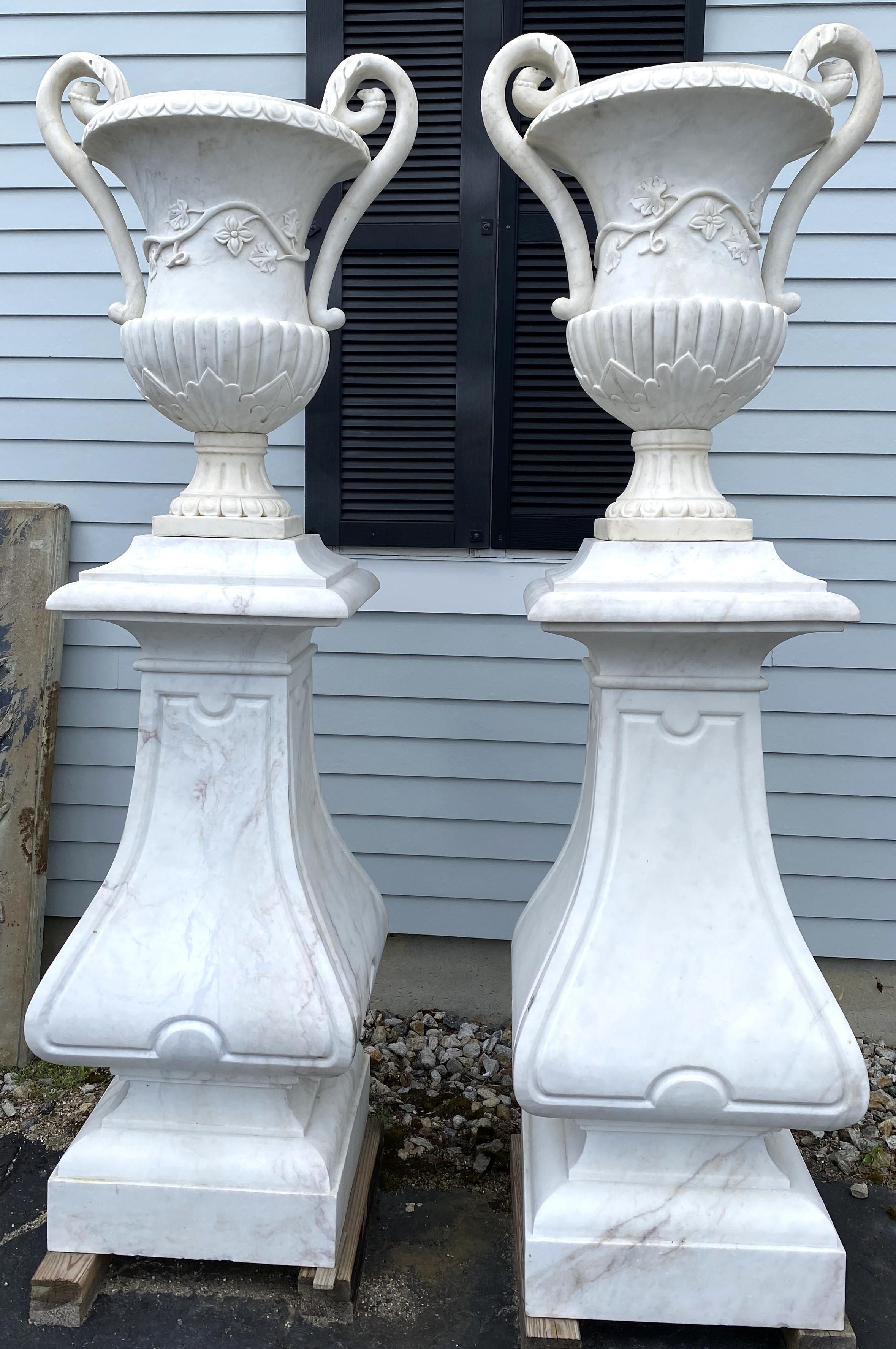 A spectacular pair of classic form carved white marble scroll-handled urns with grape leaf and vine motif, with petal edge rim design on beautifully carved marble square pedestals with shaped balusters. This pair dates to the 20th century and are in