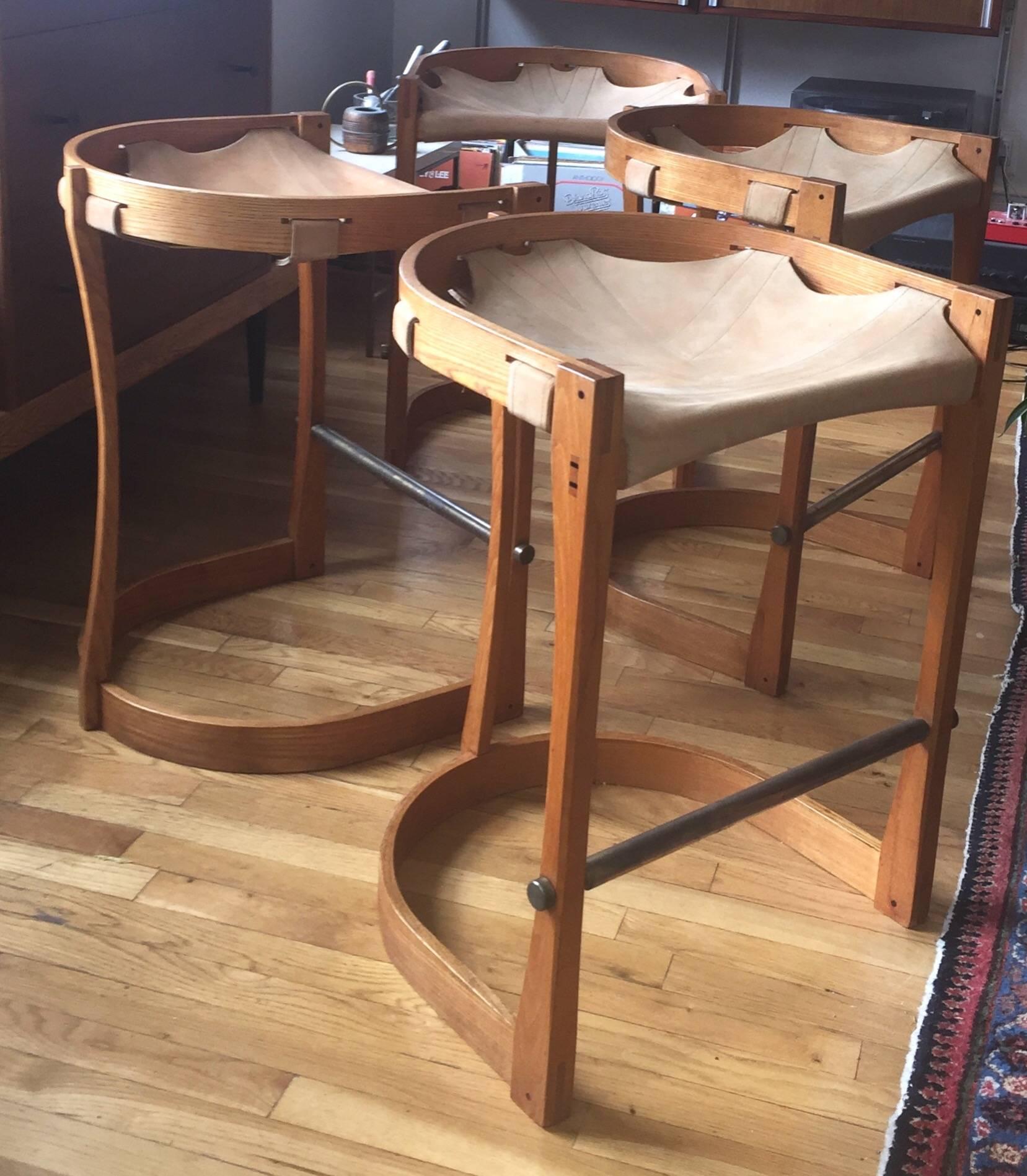 c. 1970 solid bentwood Ash stools with stitched saddle leather sling seats.  Remarkable build quality and subtle details, such as peg and dovetail construction with exposed ebony wood pegging, solid brass foot rests, and intricate  wood carving. The
