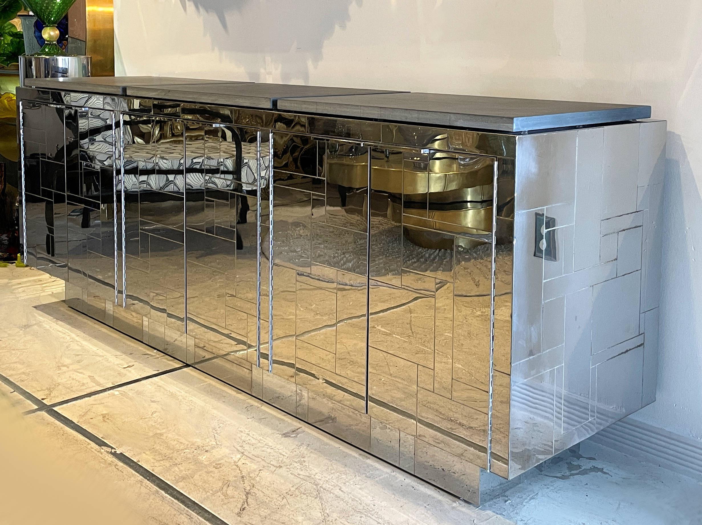 A chromium polished steel cabinet with three pairs of doors each concealing shelved interiors on either side flanking a cupboard with a single steel decorated drawer split into compartments for either cutlery or smalls. The tops are inset with