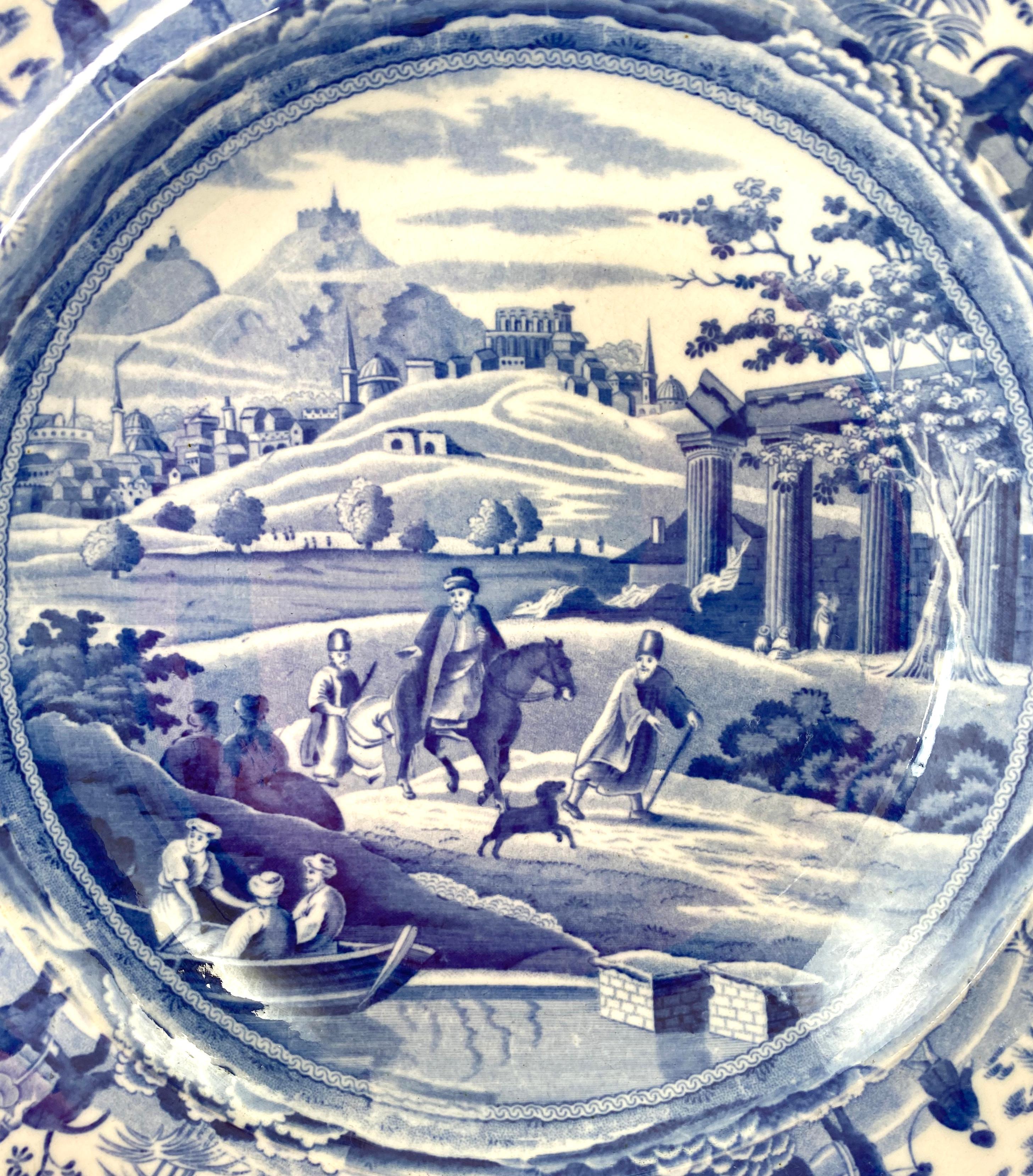 Spode Caramanian series soup plate, c. 1810. Printed in underglaze blue, with the ‘City of Corinth’; two men travelling away from the city, on horseback, whilst a colleague walks with a dog, before Classical ruins. The border with prints of animals