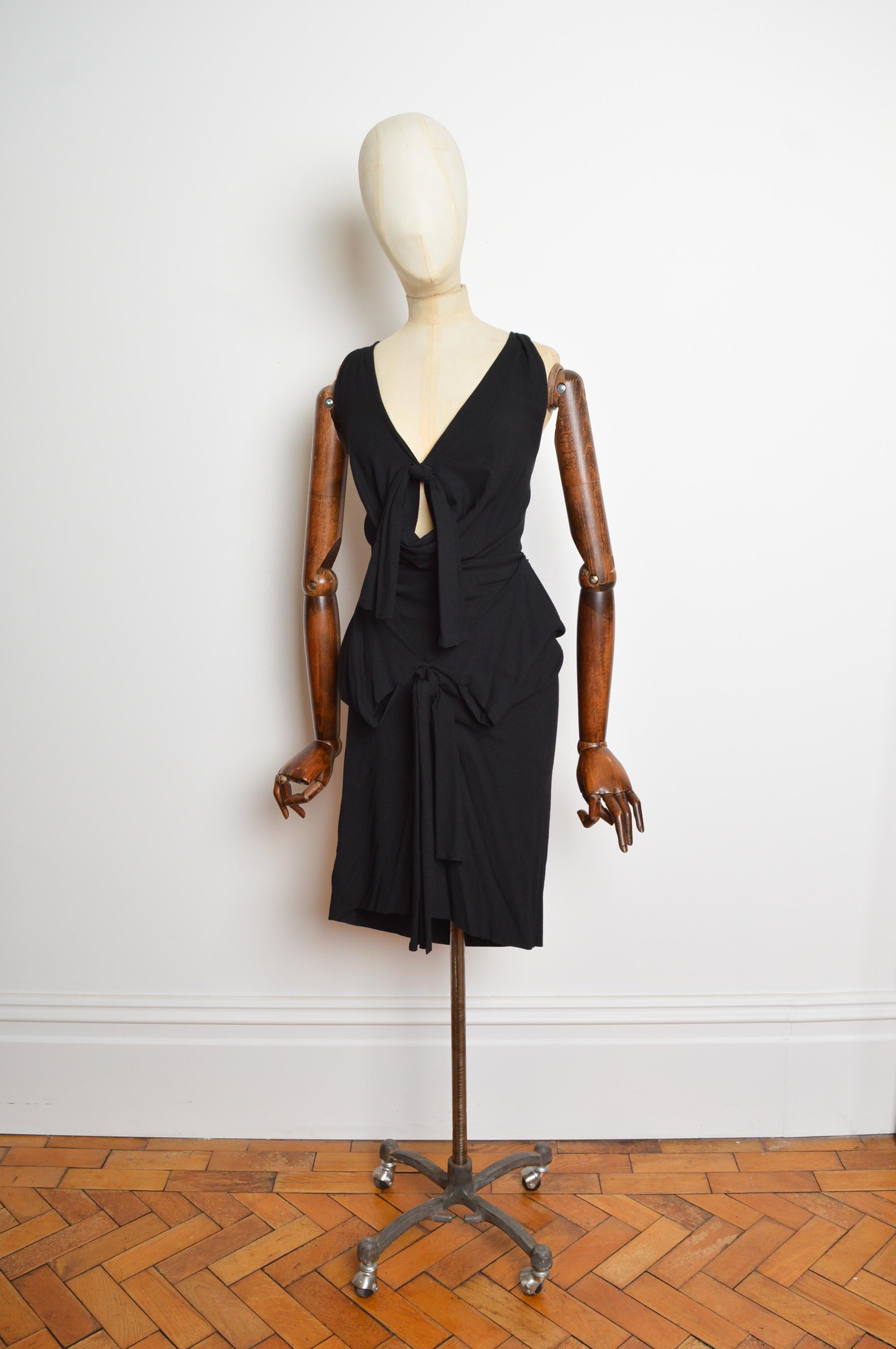 Robe de soirée noire à nœuds Bondage de Vivienne Westwood, printemps 2009, avec épaules à bretelles, encolure plongeante, taille ajustée et ourlet à mi-mollet. La robe se ferme par un zip dissimulé sur le côté.
Taille recommandée ; K.K. 10 
Les
