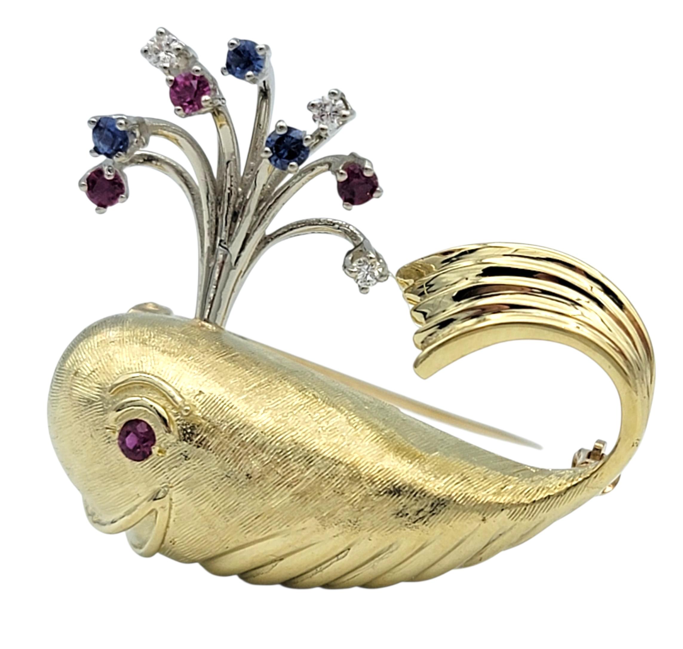 This charming 14 karat yellow gold brooch is designed in the whimsical shape of a spouting whale. The whale’s eye is set with a vibrant ruby, while its playful water spout is accented with a mix of sparkling diamonds, vivid red rubies, and rich blue