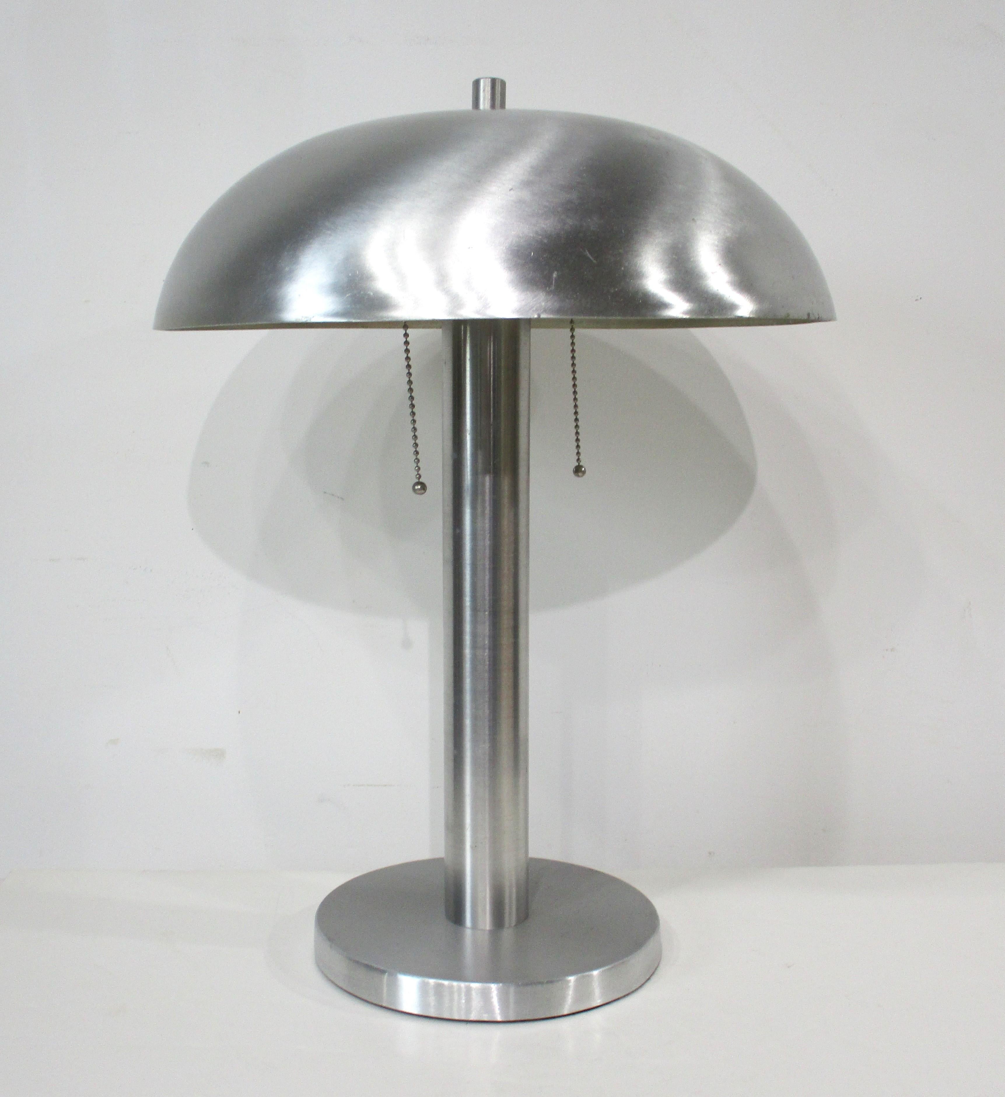Eine Art-Deco-Lampe aus gesponnenem Aluminium mit passendem gewölbten Schirm aus gesponnenem Aluminium auf einem runden Sockel mit Filz an der Unterseite zum Schutz Ihres Tisches. Eine Doppelsteckdose gibt dem Bereich, in dem sie verwendet wird,