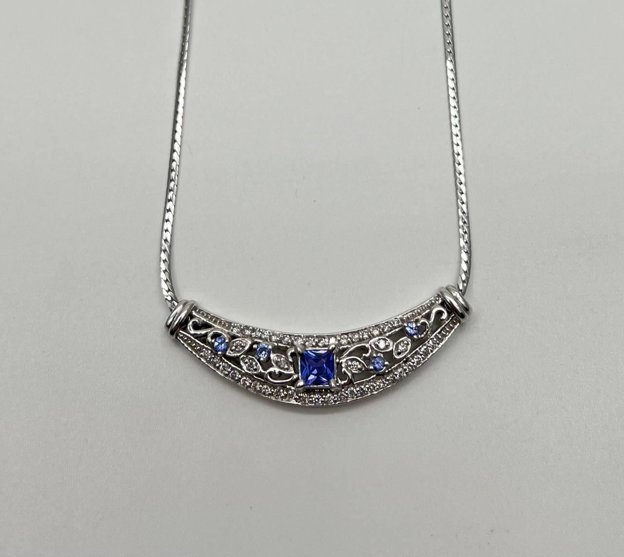 Necklace featuring a central square-cut blue tanzanite, surrounded by intricate filigree work with smaller blue sapphires and white accent diamonds. Set on a curved white gold bar with a textured chain. 14K White Gold. 0.45 CTW Diamonds. 0.66 CTW