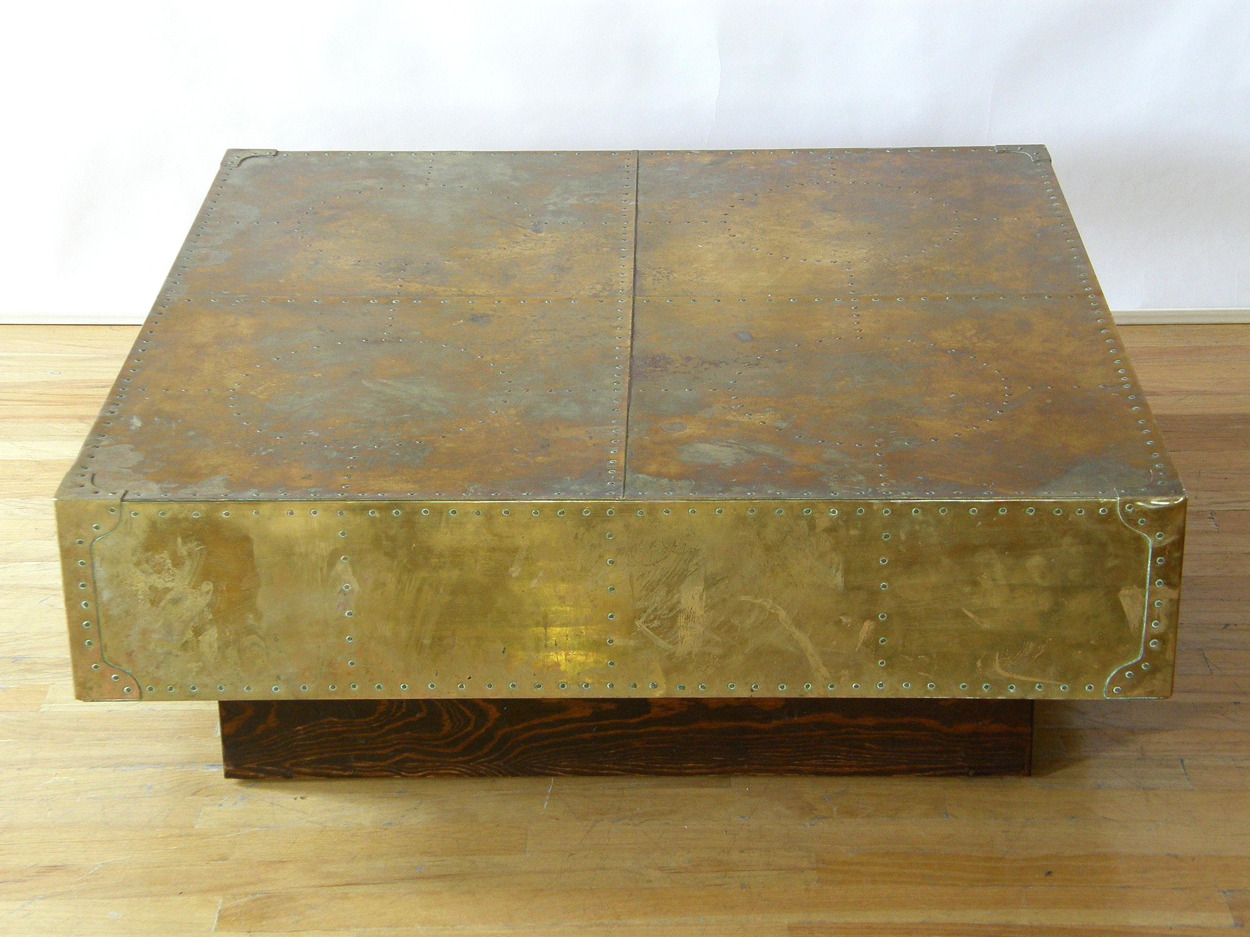 This imposing coffee table has a thick top clad in brass, and a decorative, geometric pattern done in rivets. The corners have protective covers similar to those on antique steamer trunks. The wood plinth base is inset a few inches to give the top a