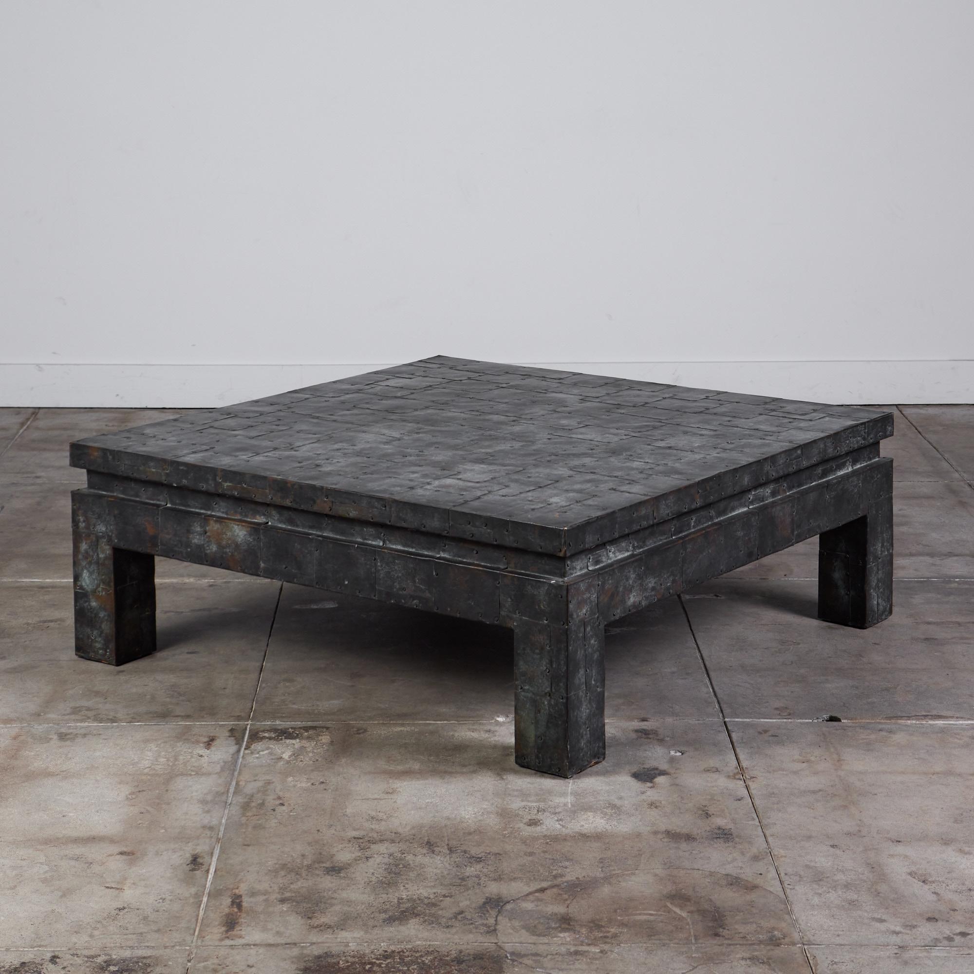 Square coffee table from North Carolina-based furniture company Maitland-Smith. The table has a wooden frame with block legs and a square body. The surface is covered with meticulously laid sheets of oxidized copper for a brutalist patchwork effect.