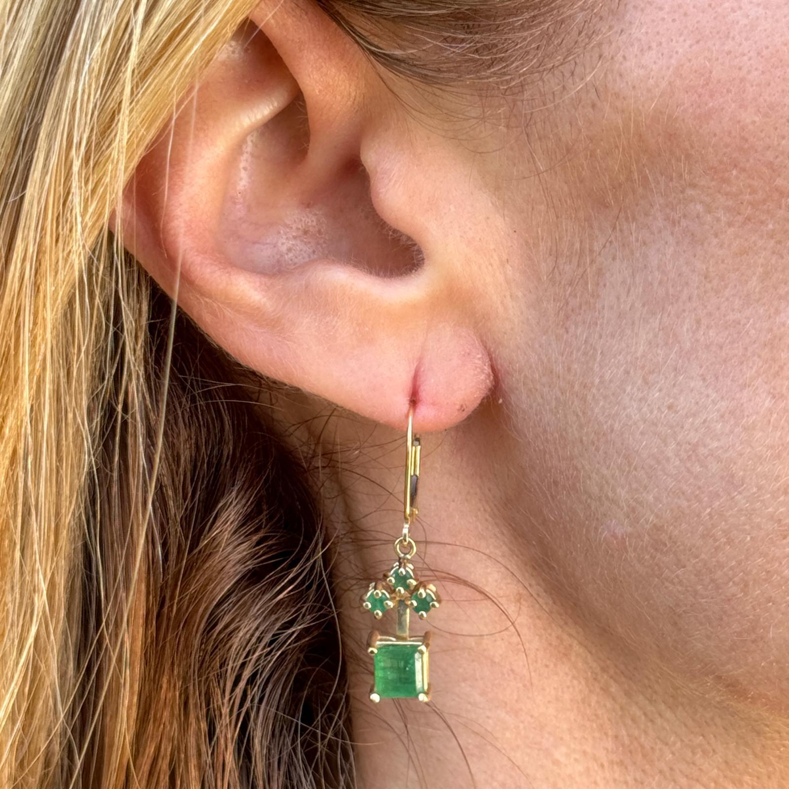 Emerald diamond drop earrings crafted in 14 karat yellow gold. The earrings feature 2 square cut natural emerald gemstones weighing approximately 1.40 CTW and 6 round emeralds weighing another .60 CTW. The earrings measure 1.30 inches in length,