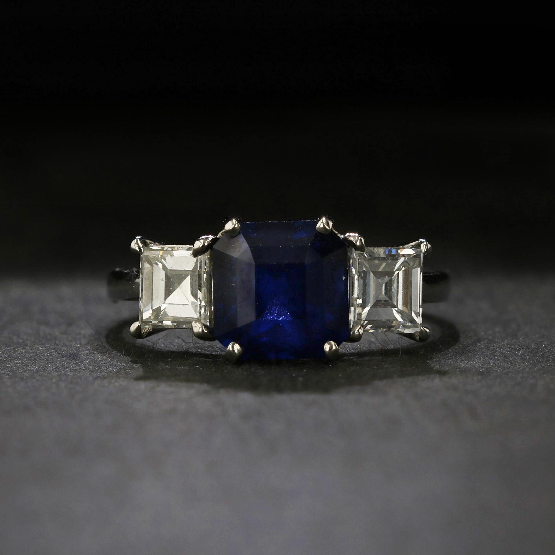 The rich royal blue of this Ceylon sapphire flashes its facets in a stunning Asscher cut, weighing a generous 3.31 carats. It is accompanied by two bright white square emerald-cut diamonds and set in gleaming platinum. The pair of diamonds together