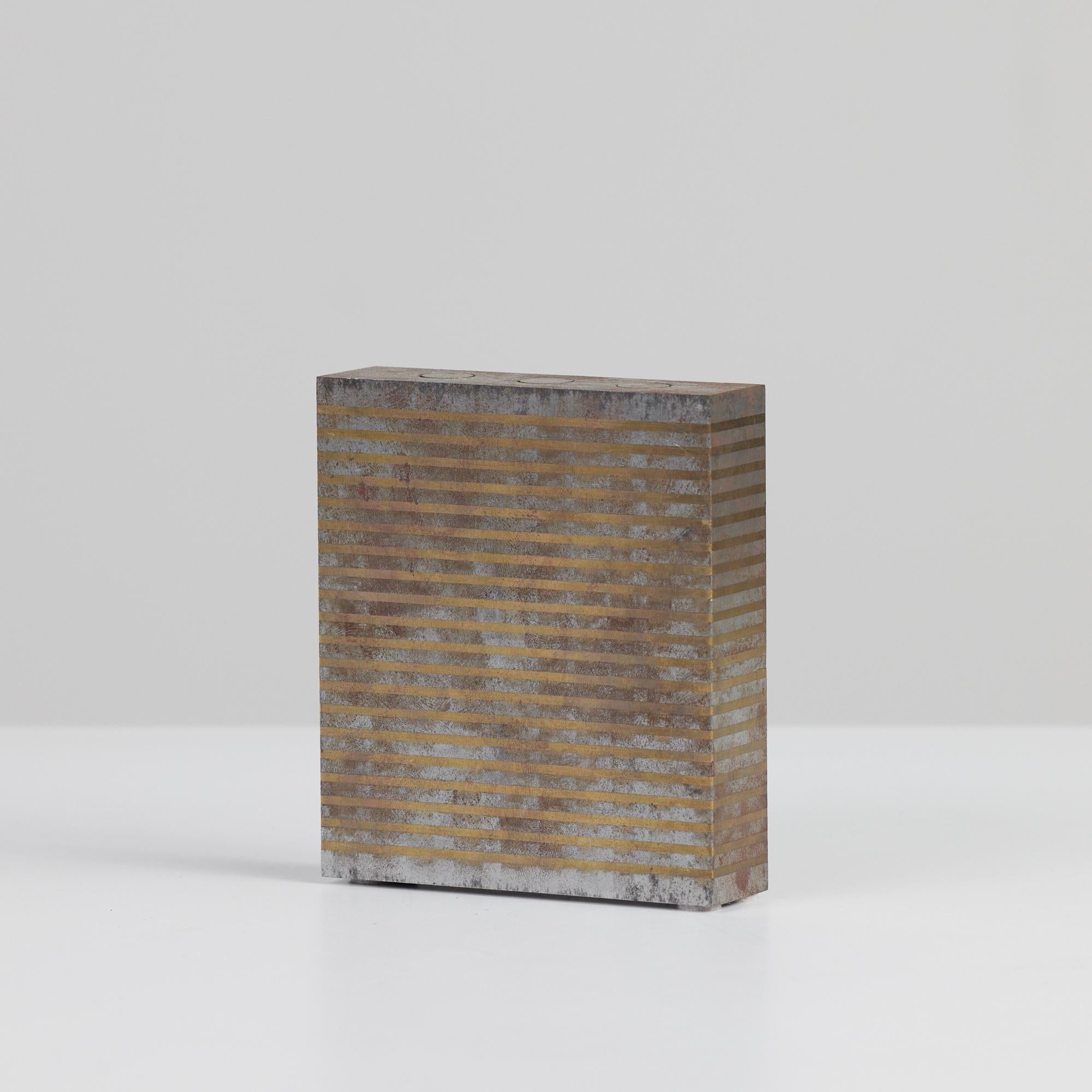 Square industrial machine block featuring alternating patinated bronze and silver pattern throughout the piece. It is flanked by three bronze inlayed 