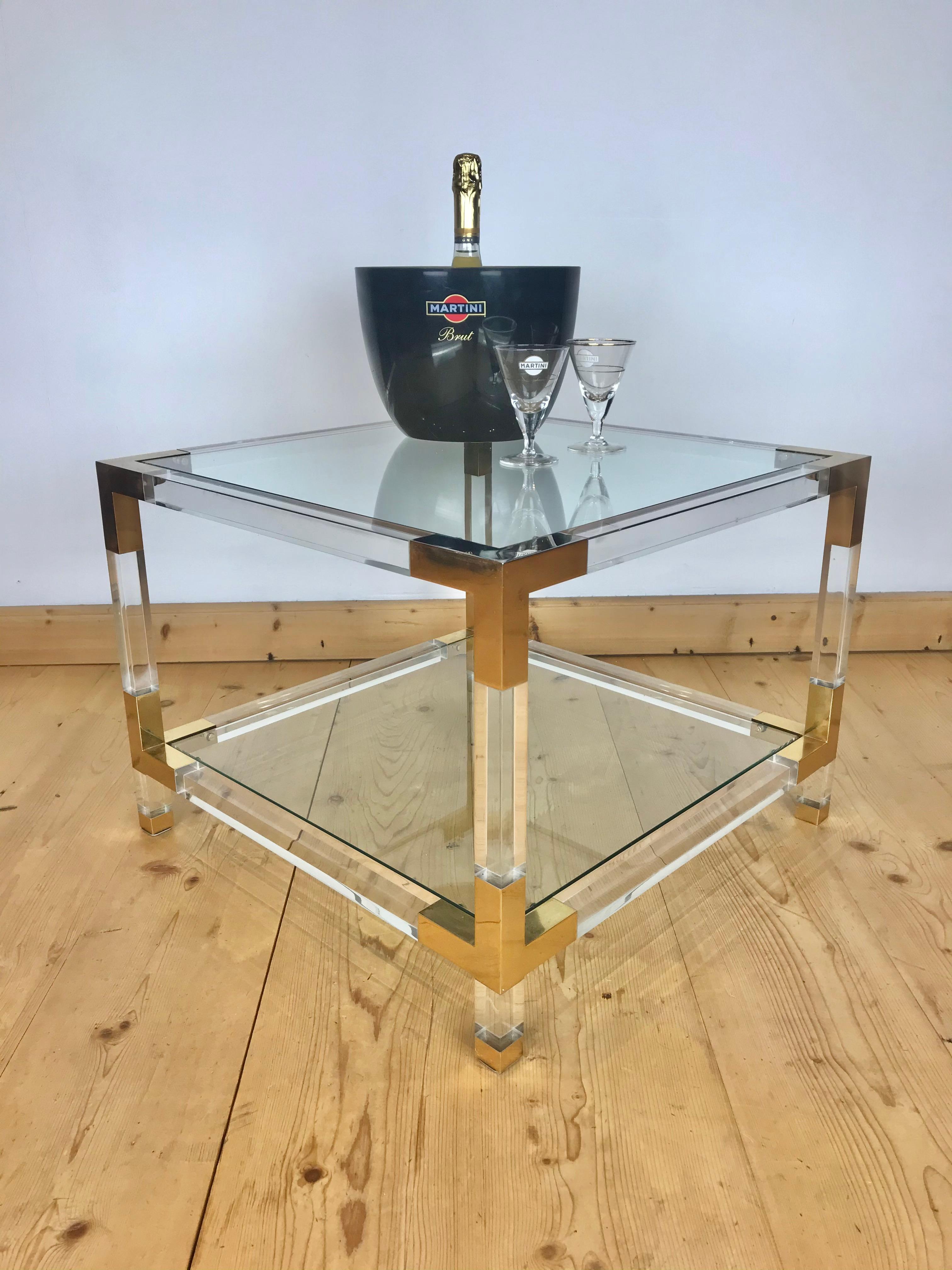 Square coffee table made of lucite, brass and glass . 
This Hollywood Regency coffee table has a lucite or acrylic base, 
polished brass details and 2 removable glass shelves.
As stylish and modern coffee table or cocktail table from the 1970s.