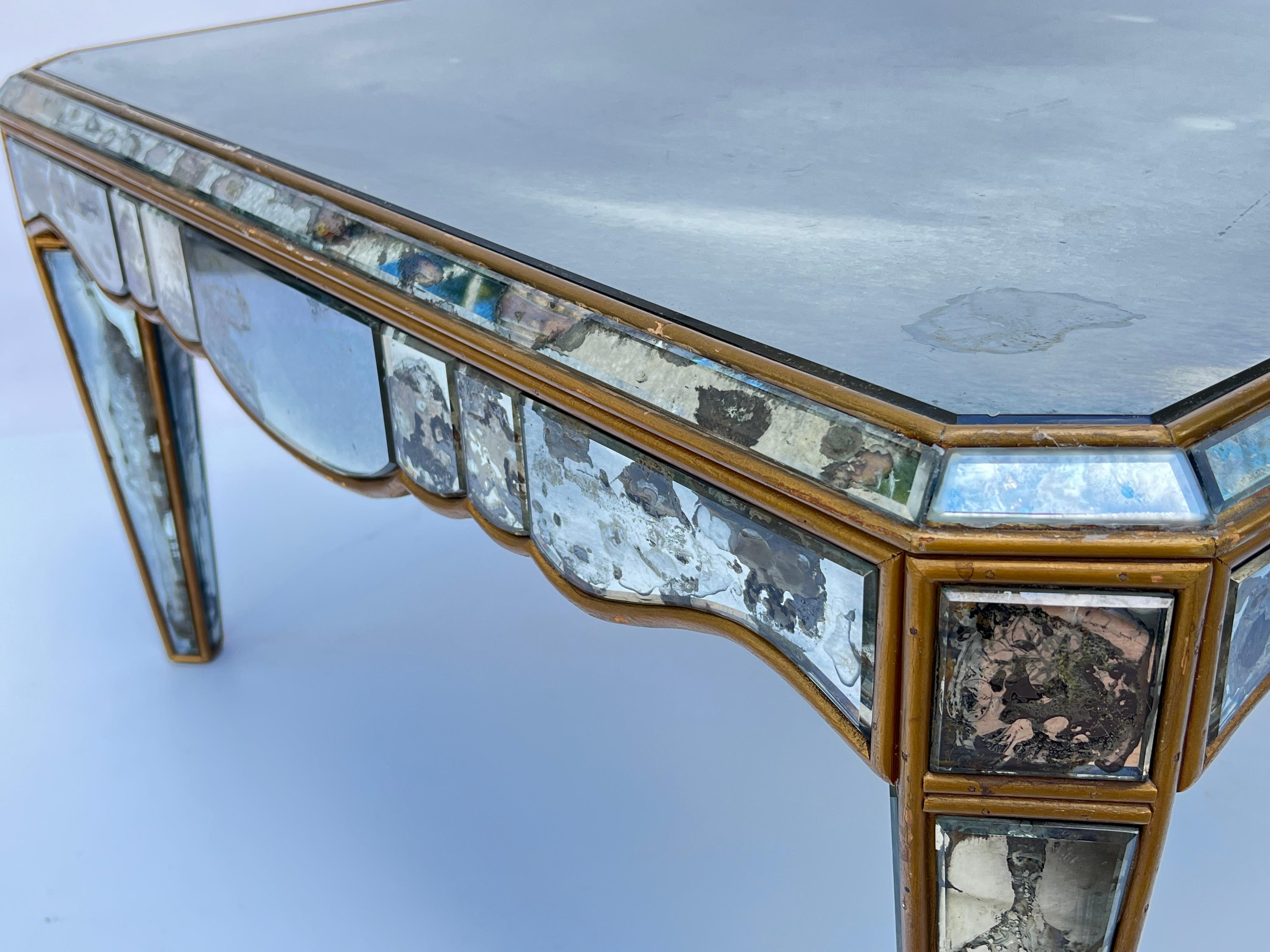 Mesa de centro de mediados de siglo, con tablero cuadrado con borde facetado y esquinas inclinadas. Su delantal festoneado se eleva sobre patas anguladas, ahusadas y cuadradas. Todas las superficies están cubiertas con el espejo ahumado biselado