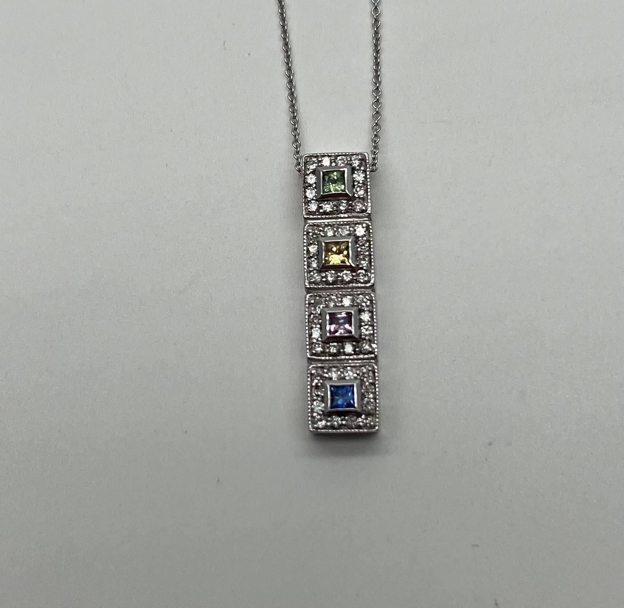 Ce superbe collier pendentif présente une disposition verticale de quatre pierres précieuses carrées, chacune étant sertie dans une délicate monture en or blanc.
Les pierres précieuses sont éclatantes, avec des teintes vertes, jaunes, violettes et
