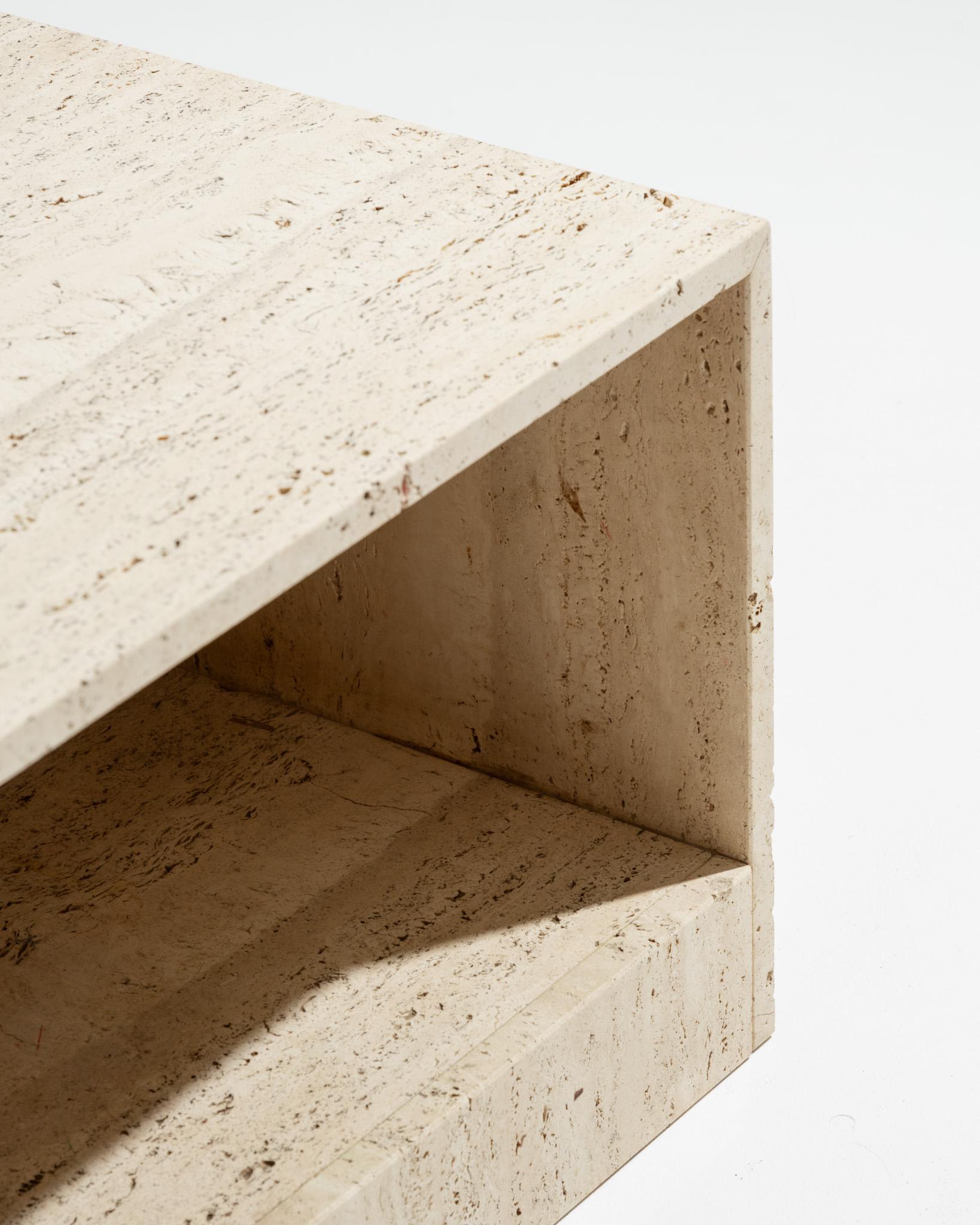 Table basse cubique en travertin des années 1970, en bloc massif, avec la partie centrale inférieure évidée formant une étagère basse, idéale pour le rangement de magazines. Elle présente un veinage naturel aux nuances de beige et est en excellent