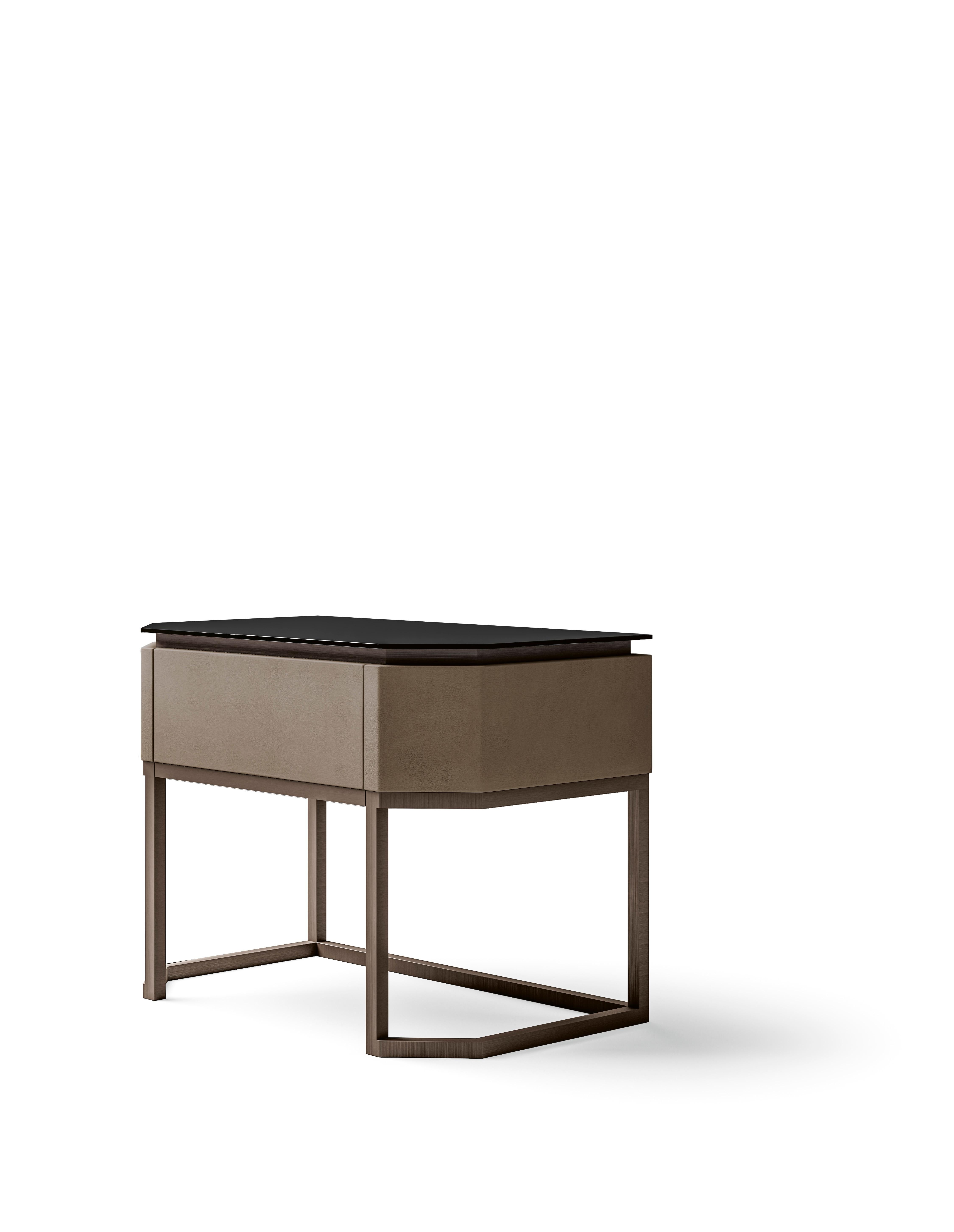 A bedside table featuring a sleek design with a smoked glass top, offering a sophisticated and modern aesthetic. The structure is elegantly crafted from high-quality leather, providing a luxurious touch, while the base is made of durable metal,
