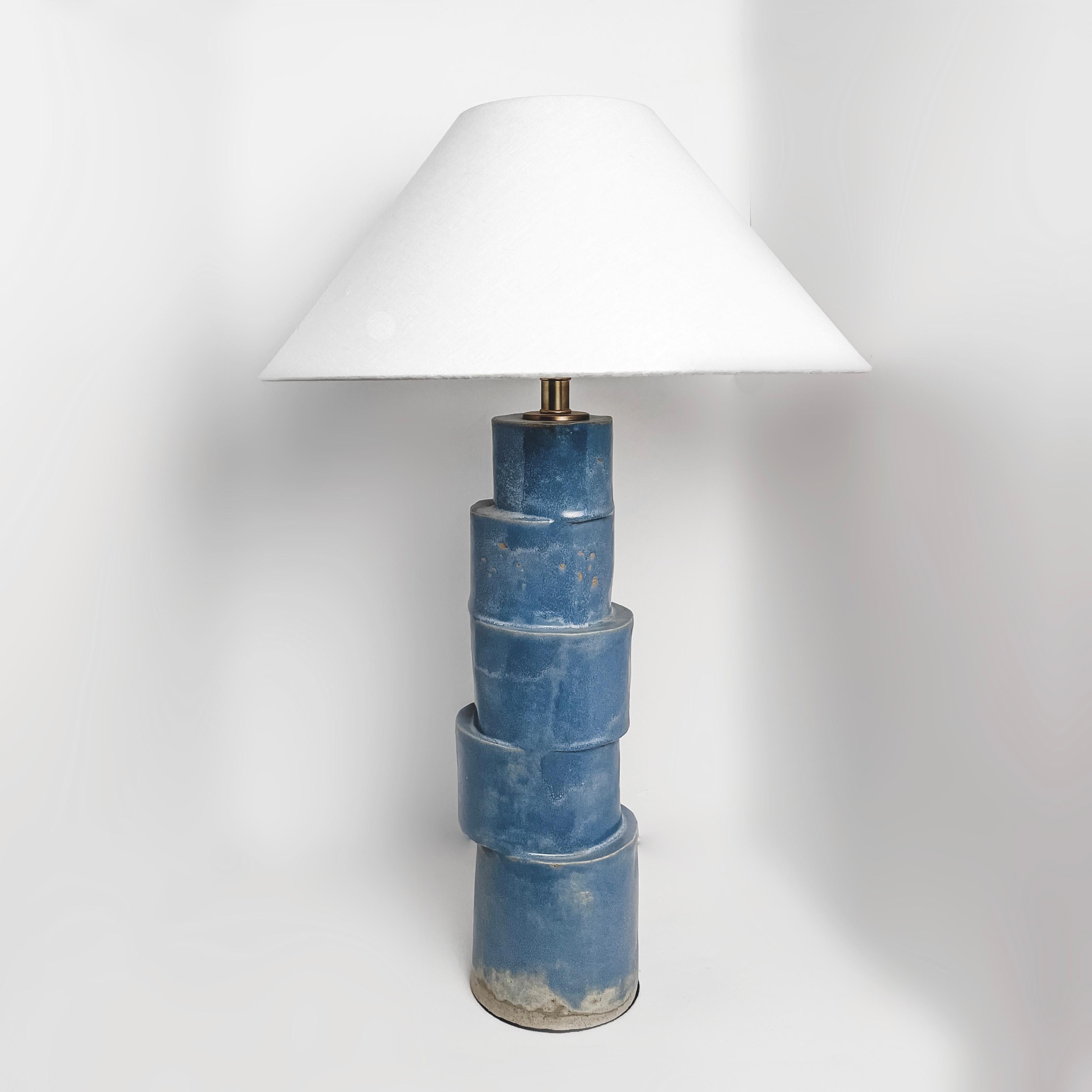 La lampe de table Stacked column est fabriquée à la main en argile raku dans une glaçure bleue avec des composants en laiton, un cordon en tissu argenté tressé et un abat-jour en lin ivoire. La nature répétitive, le ton froid et le design