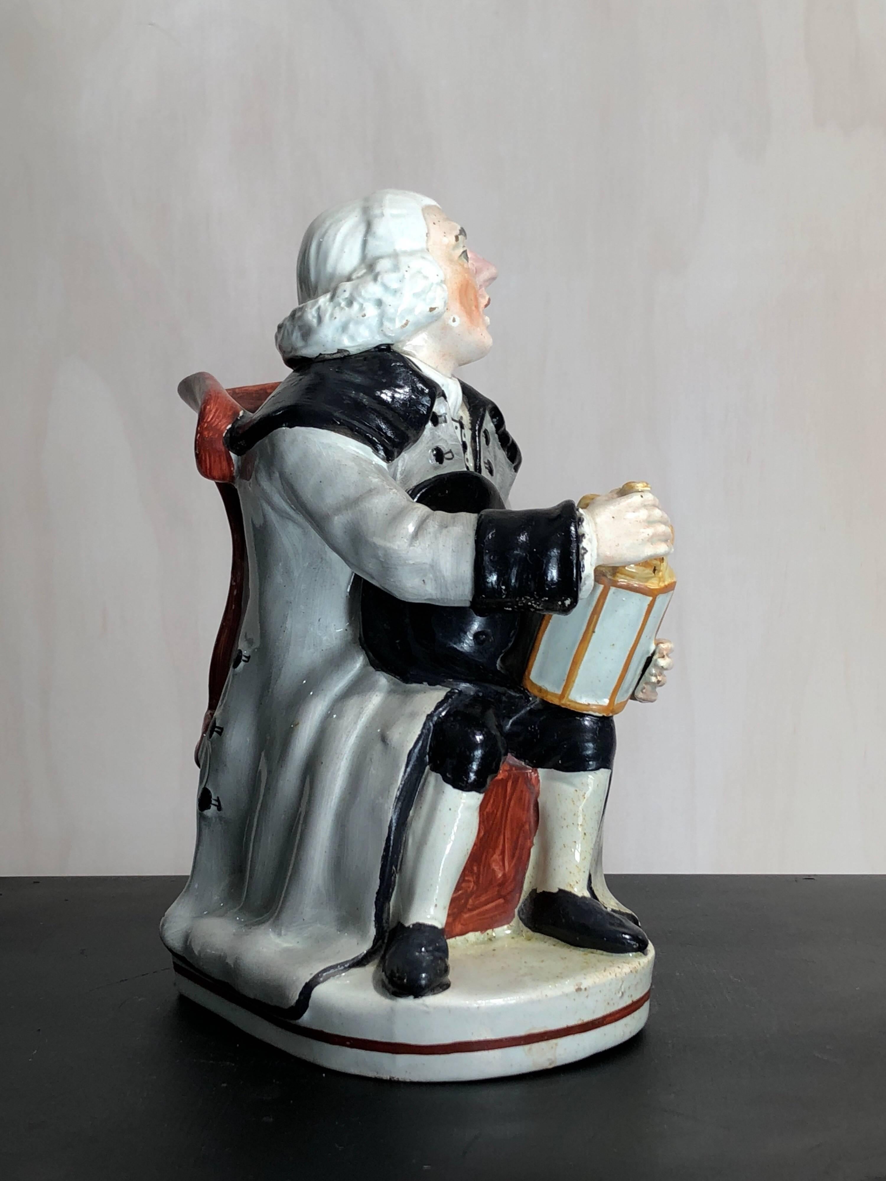 Staffordshire pottery ‘Night Watchman’ jug, formed as a man in grey greatcoat seated on a lions paw foot chair, a lantern in his hands, the back of the chair forming the spout. 
Impressed ‘5’, circa 1815.