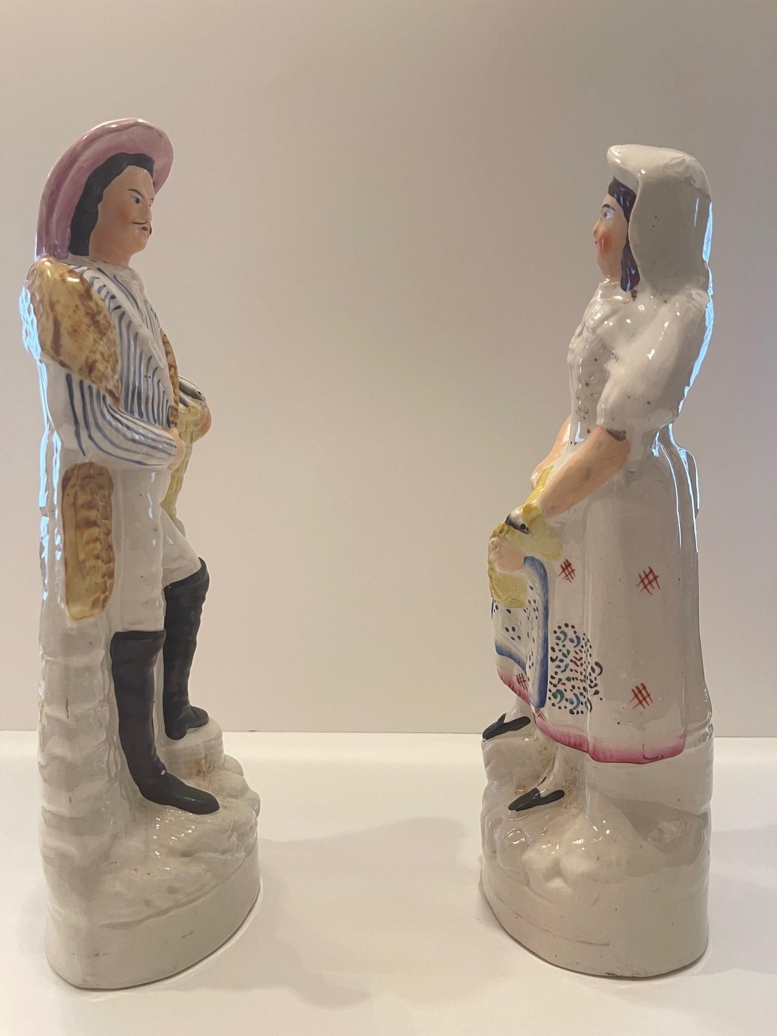 A pair of Staffordshire Figures of a woman and a man holding a catch of fish. The brown-haired lady is dressed in a peasant blouse, skirt, and apron. She is wearing a white hat and black shoes. The white tie on the hat flows into her collared white