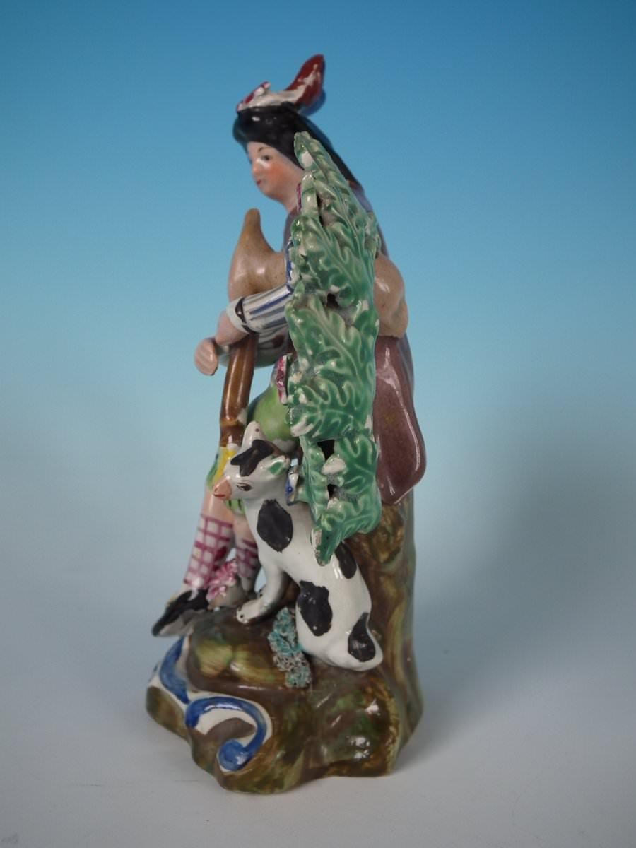 Figura di bocage in ceramica perlata Staffordshire Pottery a tema musicale con un uomo che suona una cornamusa e il suo cane accanto a lui, seduto su una base sagomata. Decorato 