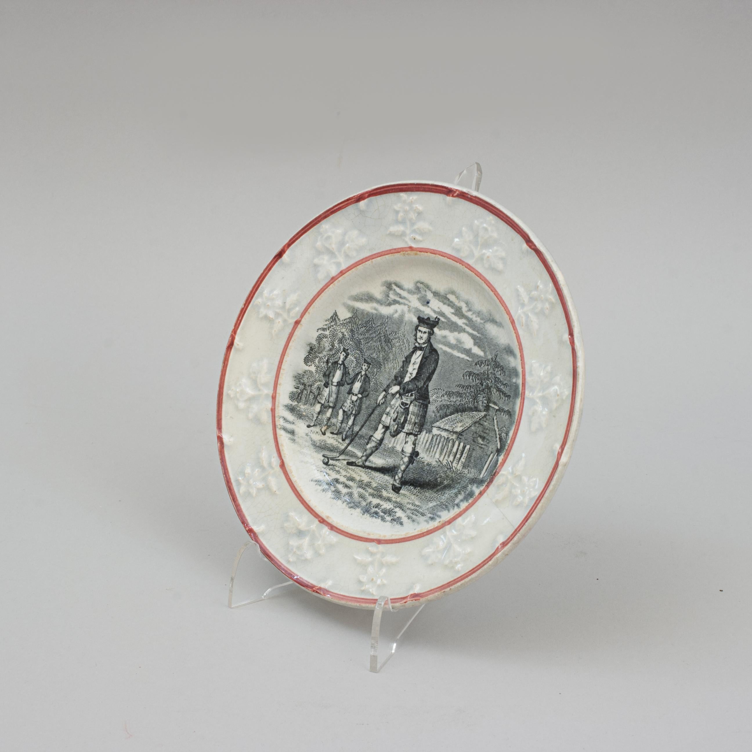 Staffordshire Ceramic Golf Plate.
An early Staffordshire transferware plate with a Scottish golf scene in the center with a flower-sprig moulded border within two hand painted red tram lines. The central scene depicts three Scottish men wearing