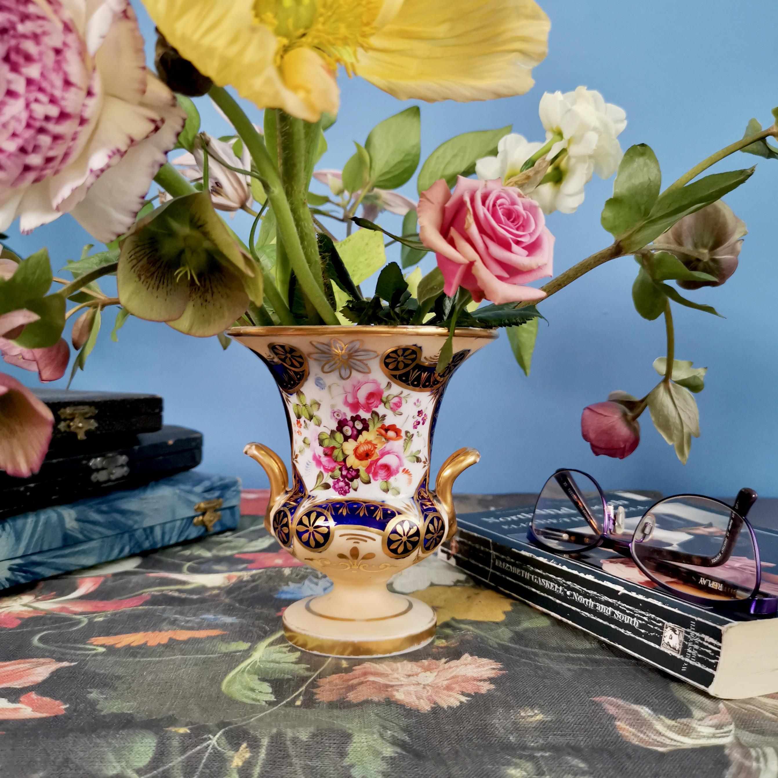 On offer is a very fine small porcelain campana vase made circa year 1820 by an unknown Staffordshire potter. The vase has a beautiful delicate salmon colored ground, rich gilding and a sublimely hand painted flower reserves on the front.

In the