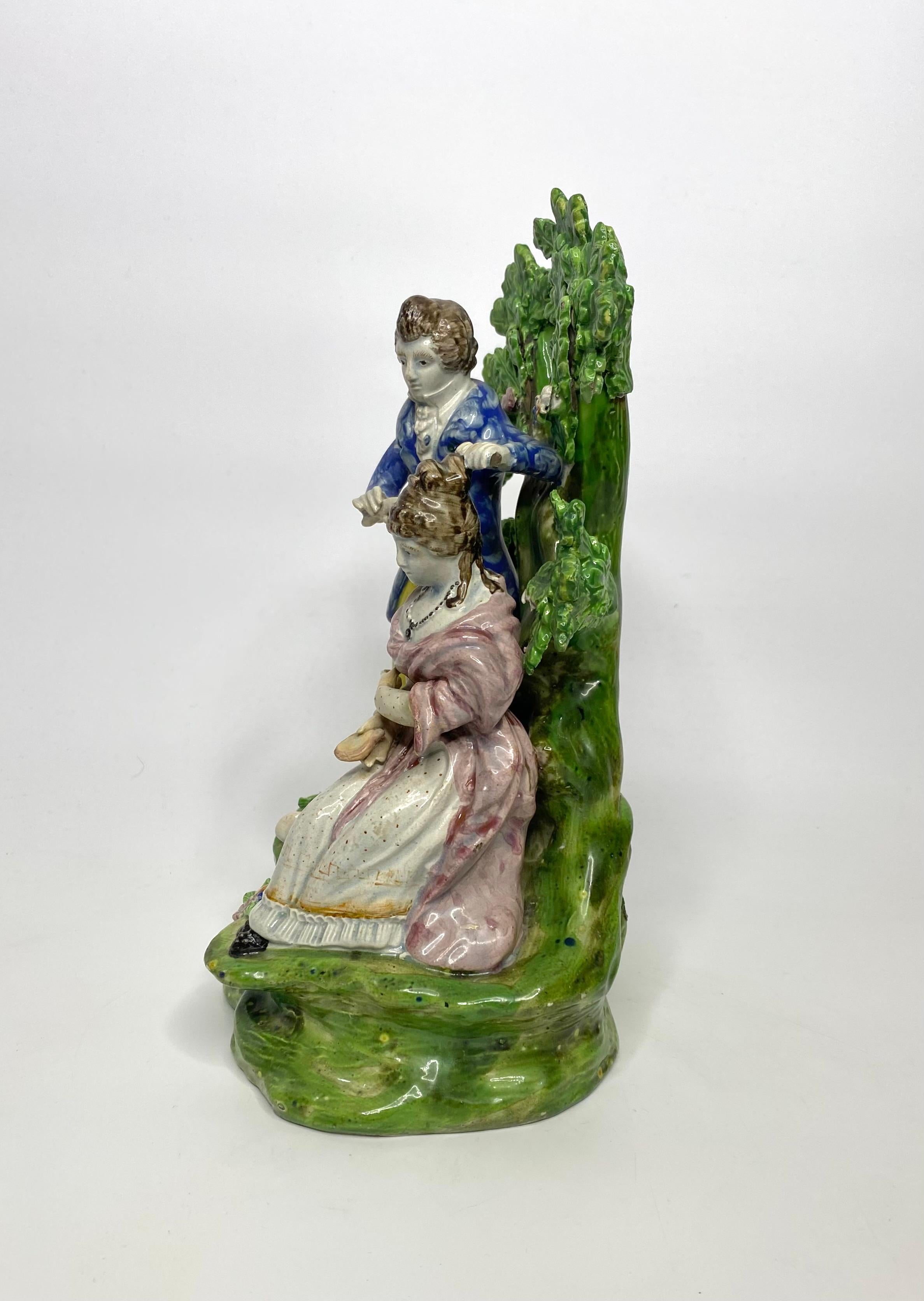 Staffordshire pottery bocage group of large size, ‘Hairdresser’, c. 1820. The very well modelled group, depicting a man curing the hair of a seated lady, whilst she looks in a hand mirror. The figures, dressed in 18th Century costume,, before an