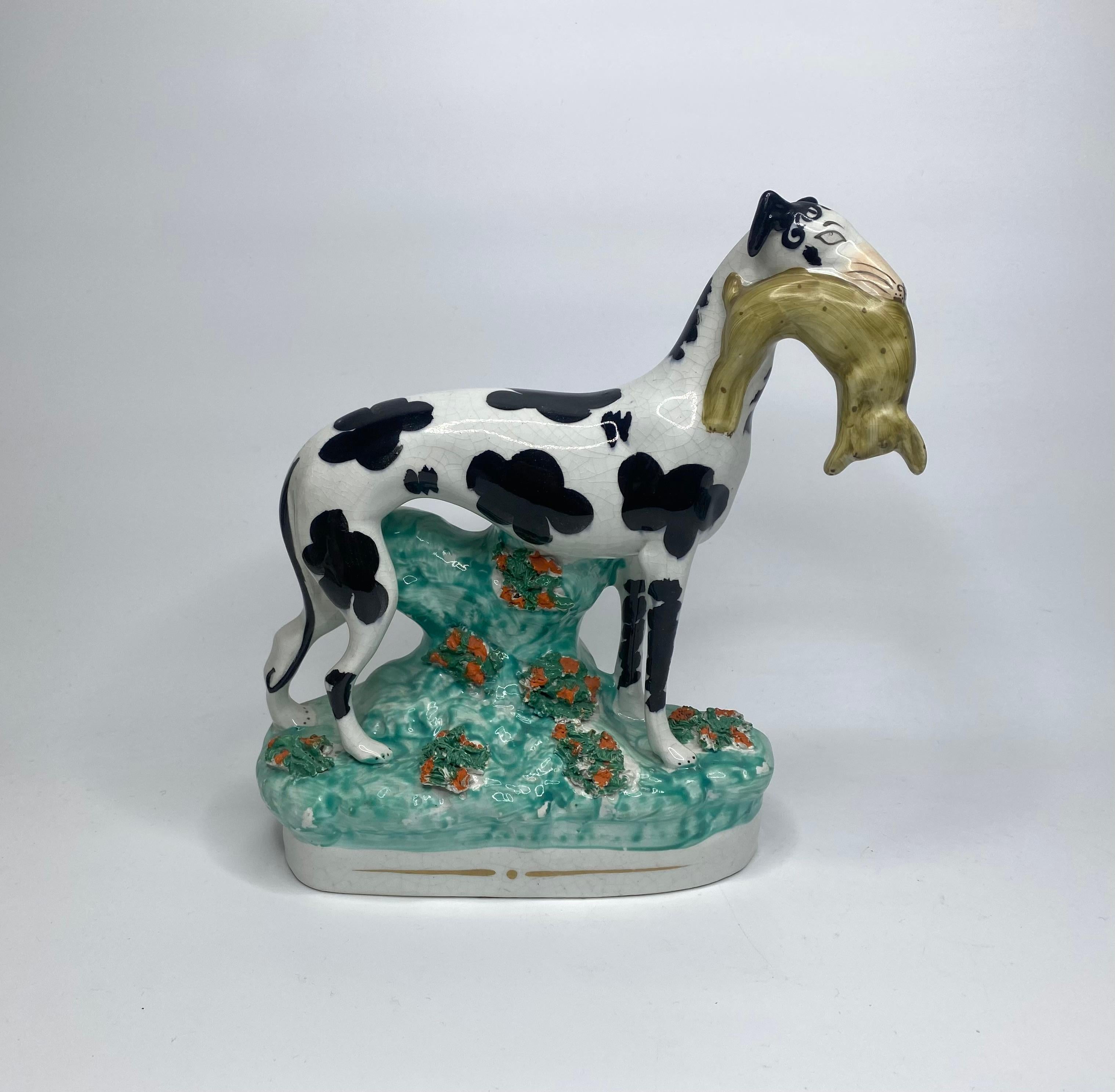 A fine pair of Staffordshire pottery Greyhounds, c. 1850. Both elegantly modelled hounds painted with large underglaze black spots, and ‘Disraeli Curls’ to their heads, and are stood with their catch of hares. Set upon grassy mound bases, with a