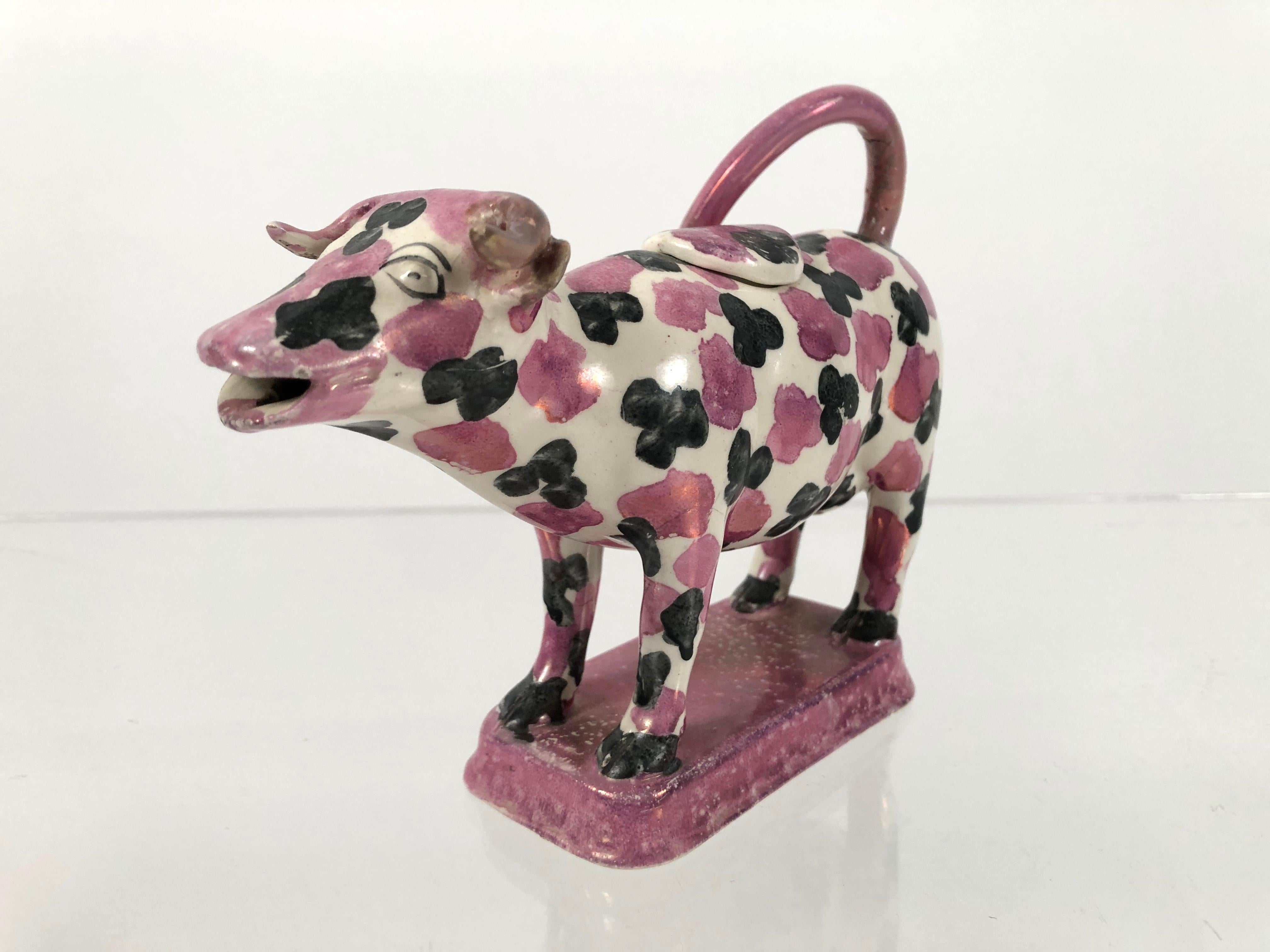 A Staffordshire pottery pink lustreware cow creamer and cover, with wonderful black and pink spotted decoration overall. The cow has an endearing happy expression and is standing on a rectangular pink lustre glazed plinth, English, circa 1820.