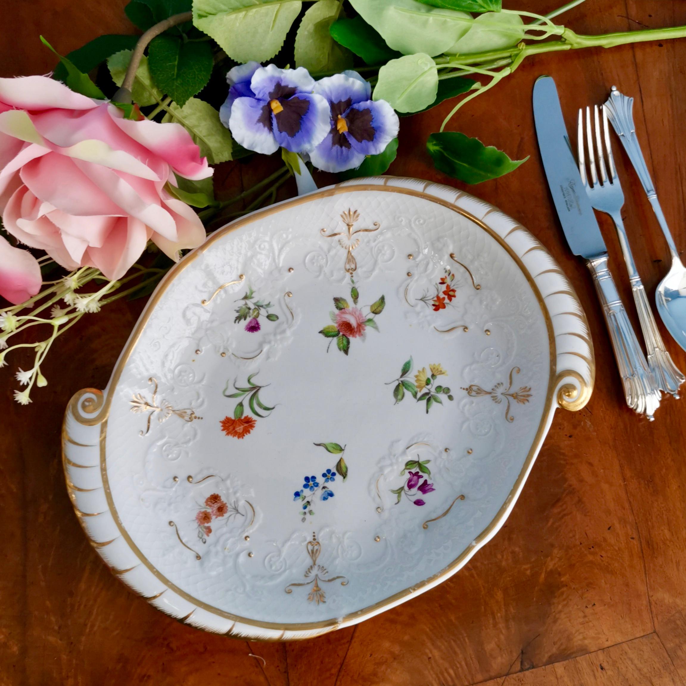 This is a very fine and beautiful serving dish made in Staffordshire in the very early 19th century. The dish has a striking oval shape with gilded handles, superbly fine surface moulding and very skilfully painted wild flowers dotted over the