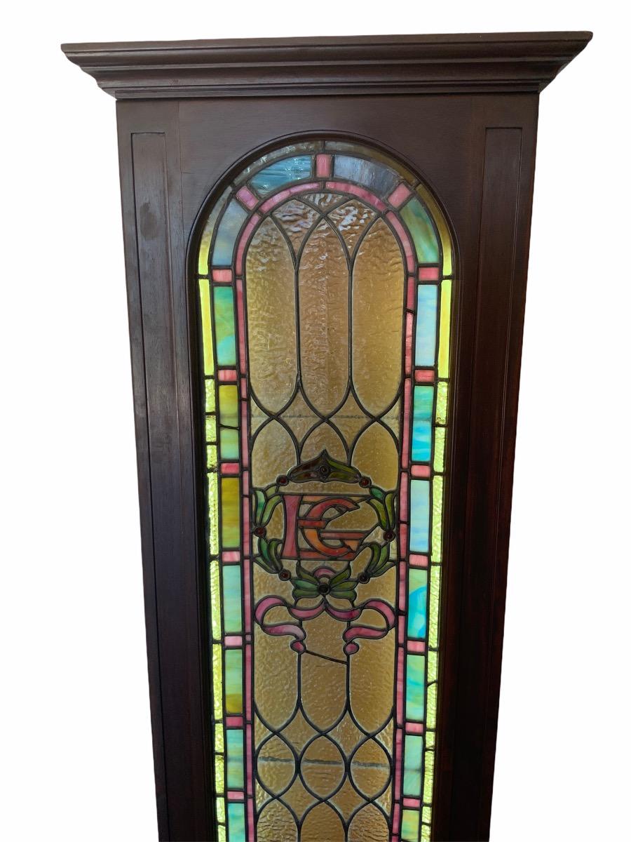 An old stained glass window designed as a bell shaped arc that is decorated in the center with a green leave wreath with flirtatious pink ribbons highlighting the letters EG of its former owner. A combination of rolled and slag polychrome glass