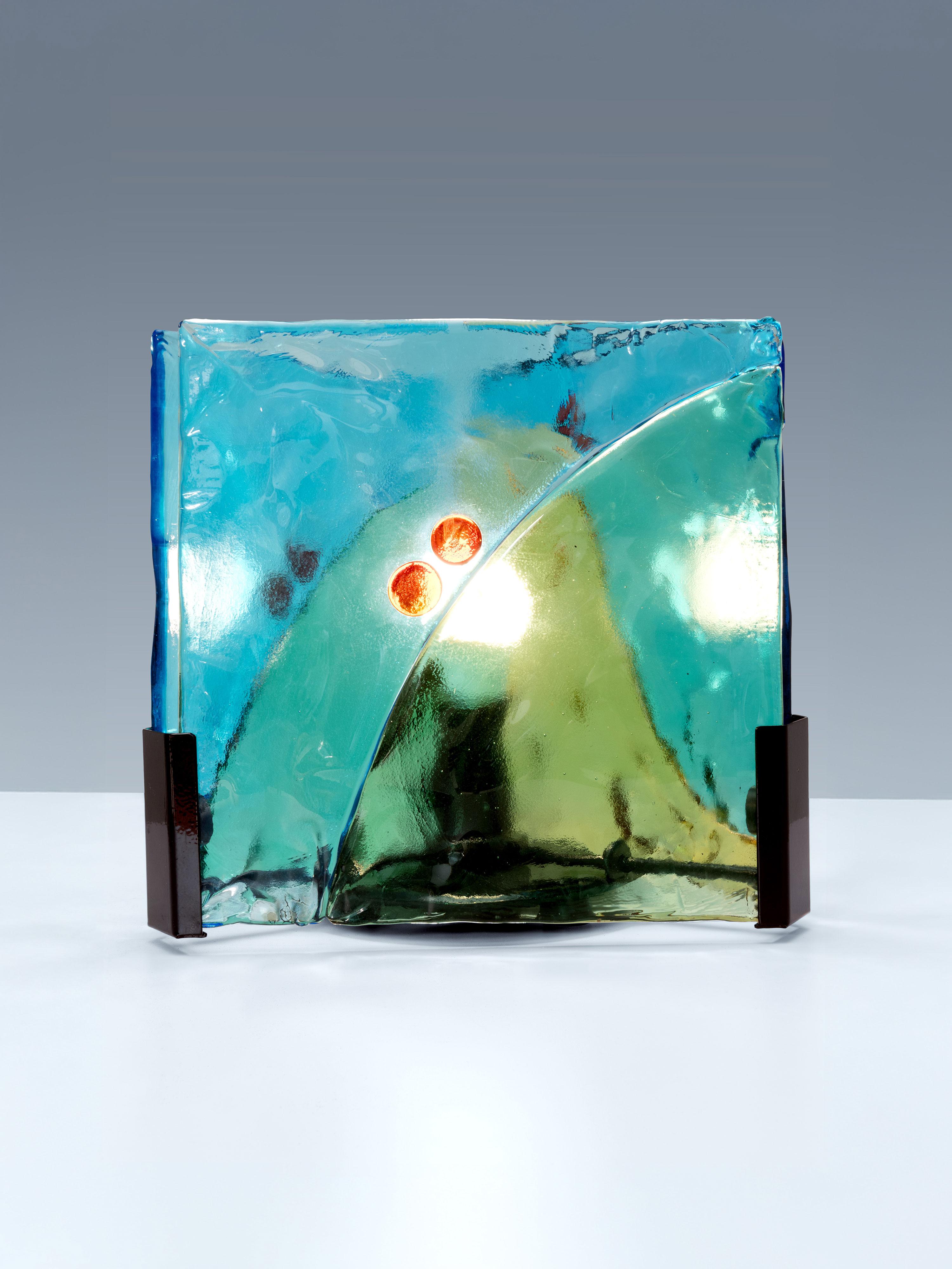 Here we have a table lamp in stained Murano glass. It consists out of three glass panels, attached to the base by a metal rail, which makes the lamp rotatable. The panels are positioned in a triangular configuration. 
These panels display a wide