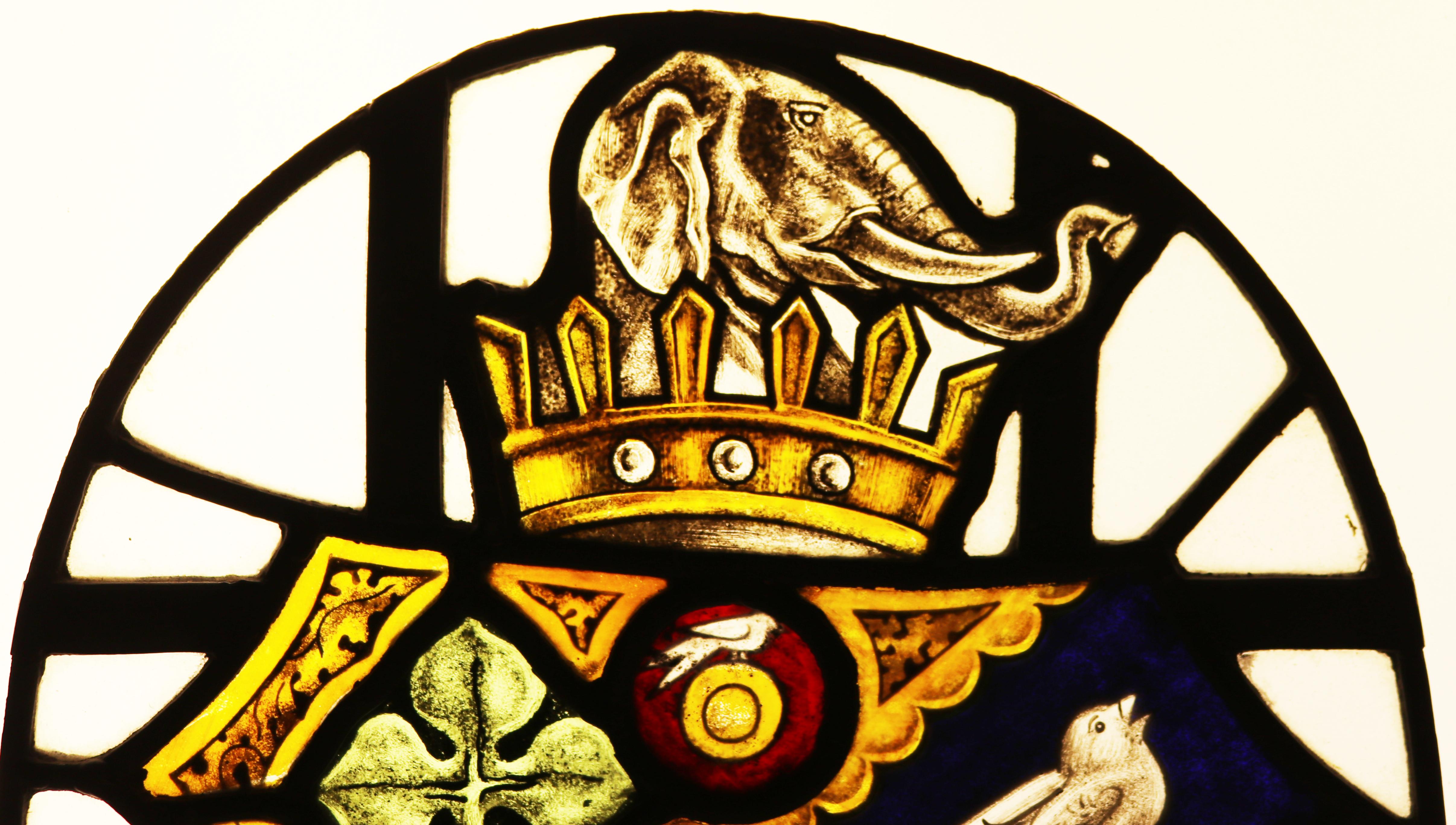 A stained glass window panel depicting a coat of arms, featuring a crown, elephant, song birds and clover. Originally fitted to a door.