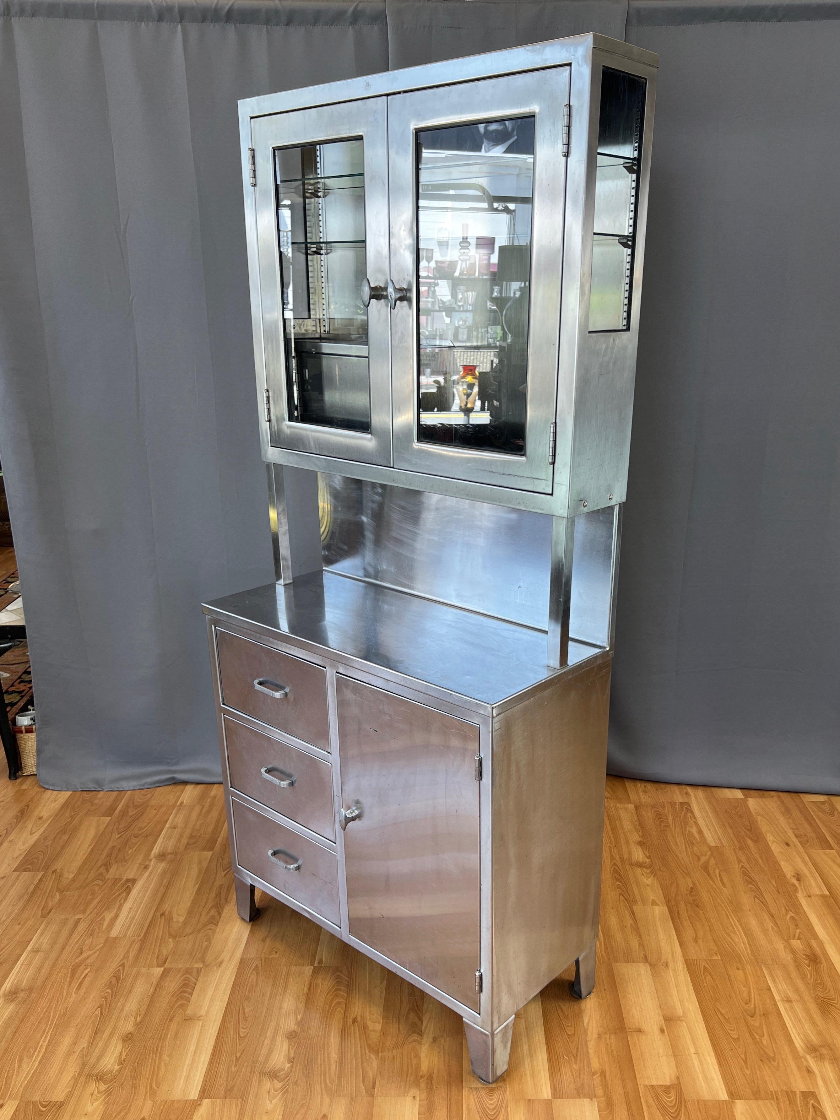 A tall and substantial circa 1950s medical or laboratory stainless steel hutch with glass doors, cabinets, and drawers.

Upper portion features a display cabinet with glass pane doors and sides, inside which are two adjustable-height glass shelves