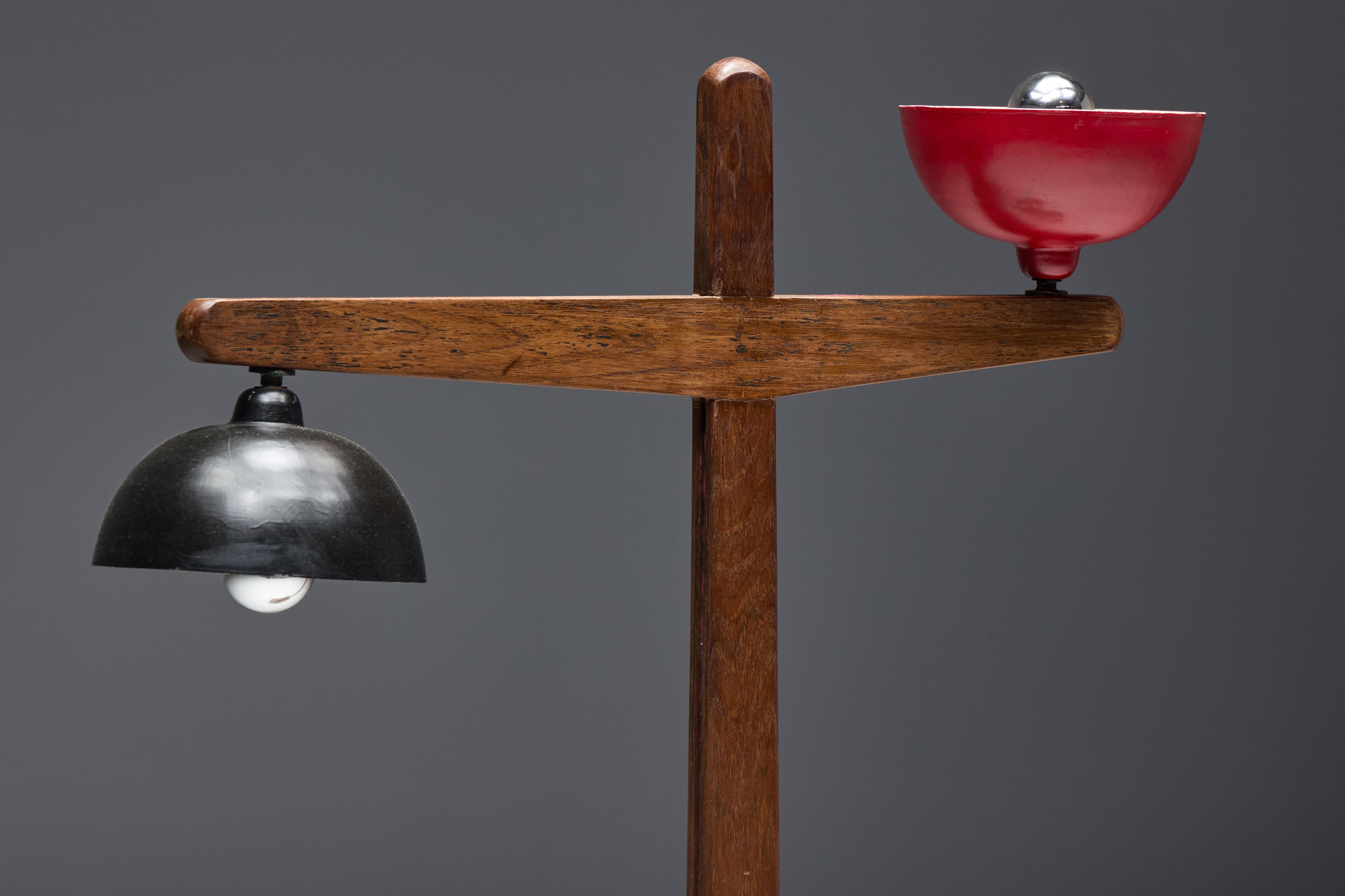Crafted in solid teak, this iconic 1955 floor lamp by Pierre Jeanneret features two dome-shaped lights in embossed aluminum, lacquered in black and red. The tapered central axis intersects with a horizontal arm supporting the lights at each end,