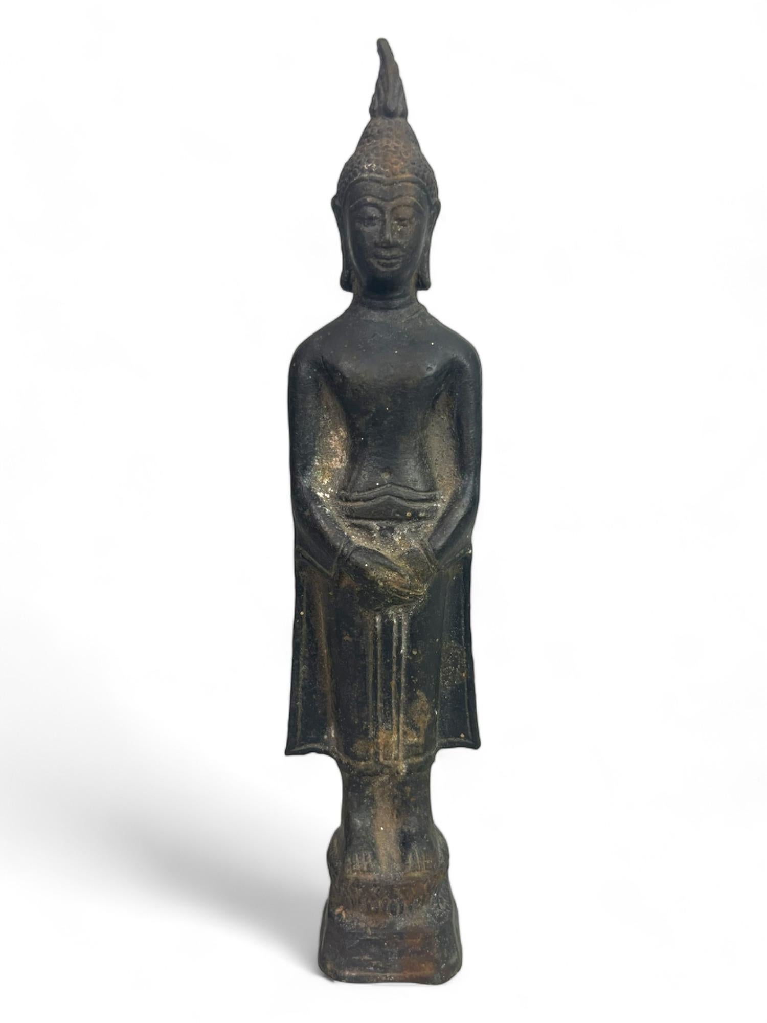 Graziosa scultura in bronzo che rappresenta il Buddha in piedi, con le mani delicatamente incrociate davanti al corpo in un gesto di meditazione (samādhi mudrā).
Il volto sereno, coronato da una fiamma allungata (ushnisha), riflette il risveglio