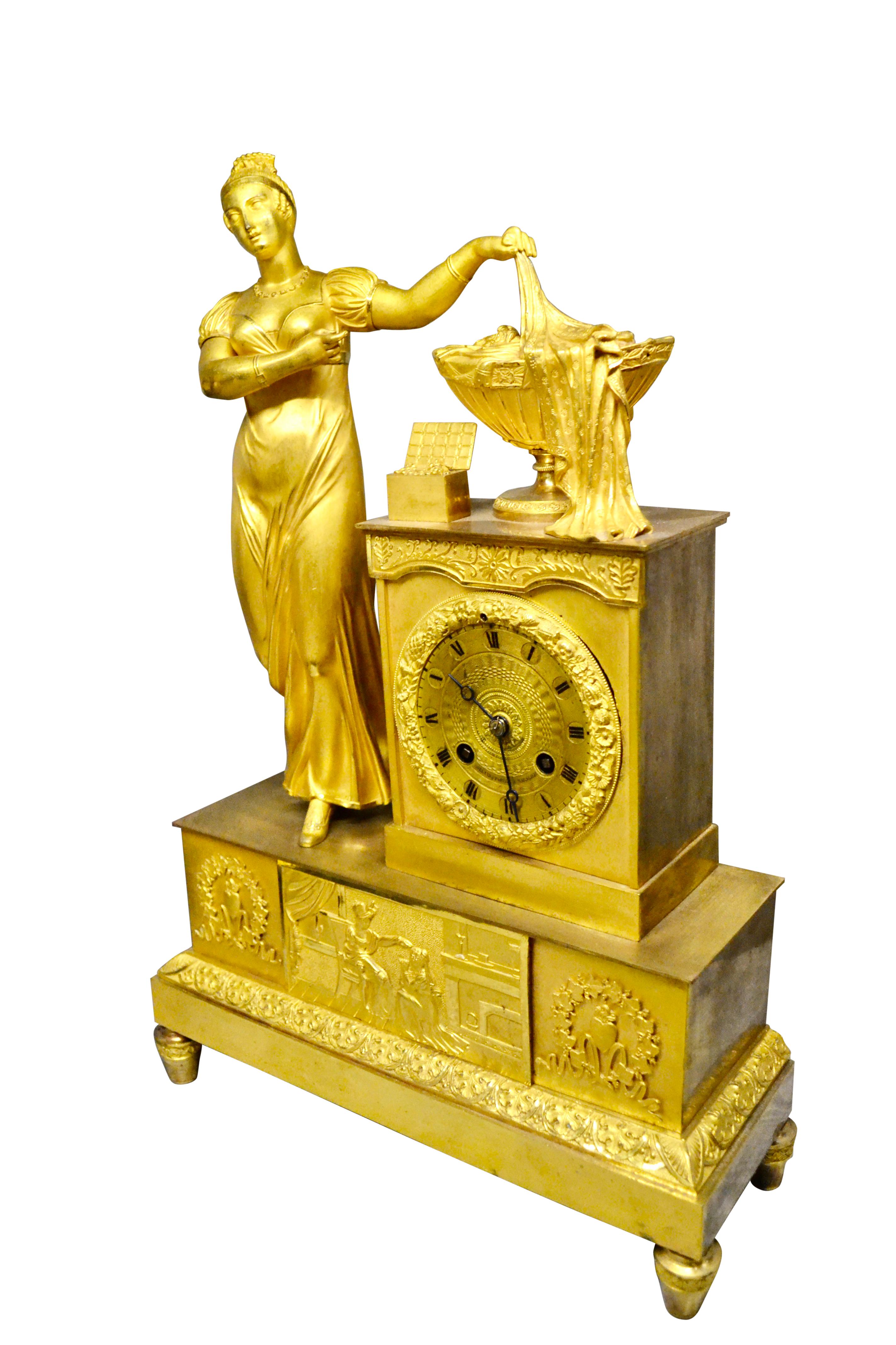 A late French Empire mantle clock in gilded bronze depicting a standing classical lady on the left side of the clock plinth with her left arm outstretched holding drapery concealing the contents of a gilded urn. The urn contains numerous items
