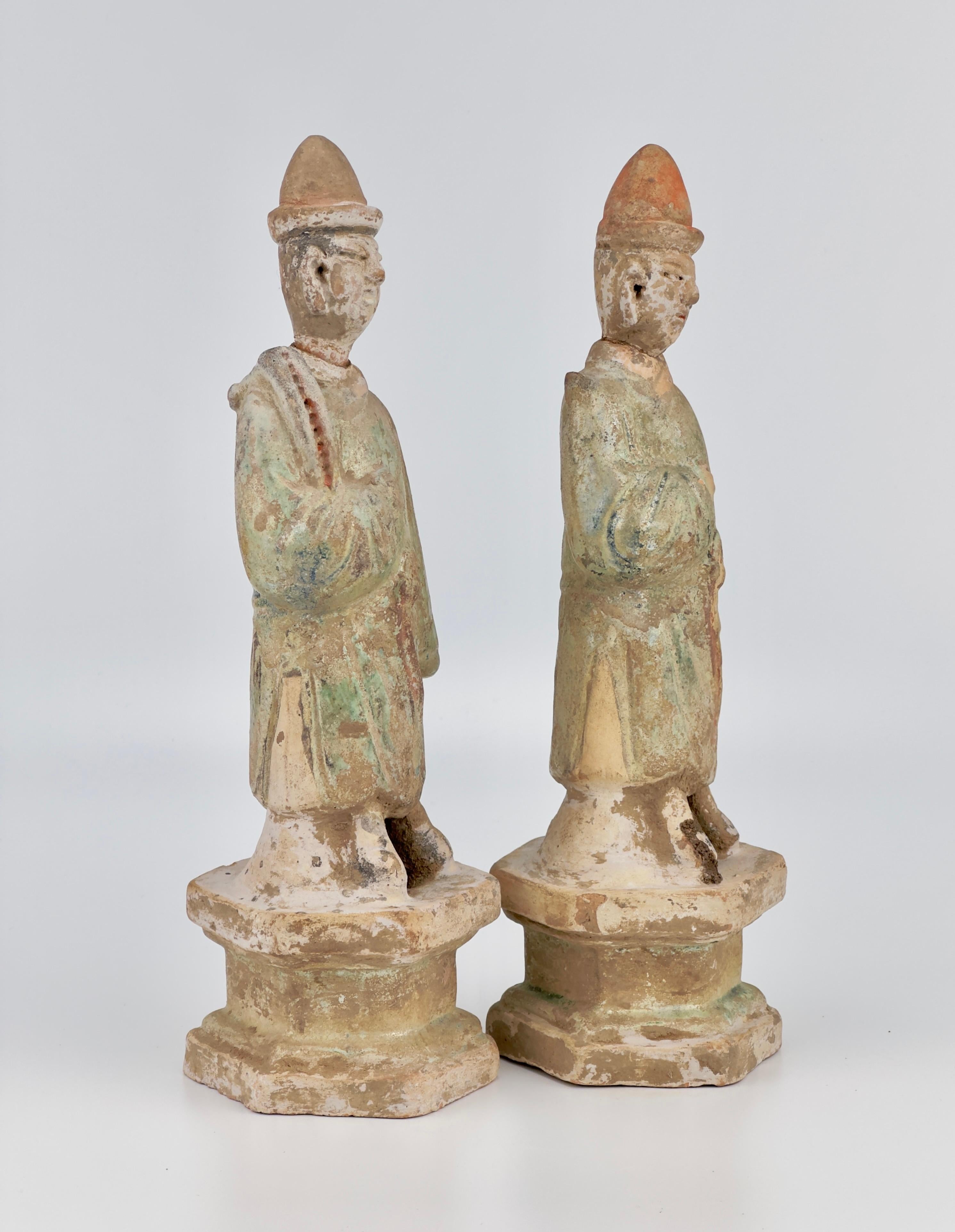 This standing attendant male figure is dressed in a front-closing green robe, cinched at the waist with a sash tied in a knot. Atop his head sits a red hat. He adopts a formal pose on a hexagonal stand, his right hand crossing his chest. The green