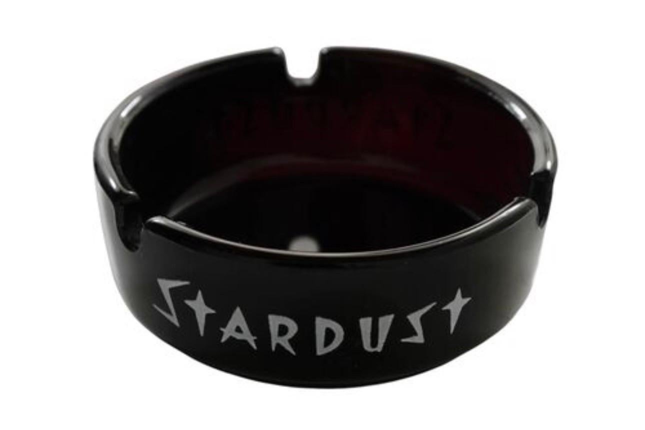 Ein schwarzer Glasaschenbecher aus dem Stardust Hotel & Casino in Las Vegas, Nevada. Der Rand ist in Weiß mit dem Stardust-Logo bedruckt.

