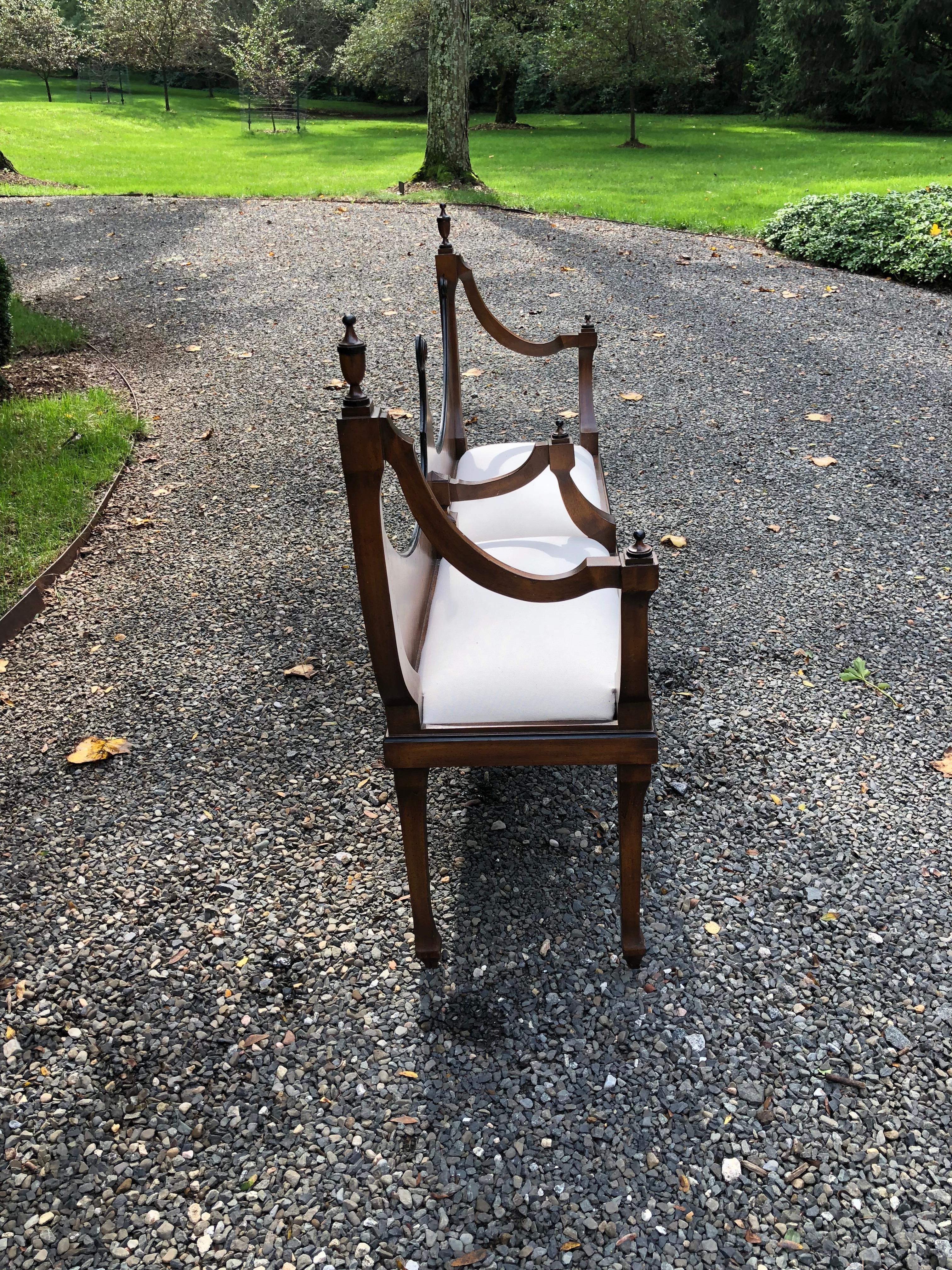 Stately neoclassical style fruitwood bench having an inverted double arched back. The back of the bench is decorated with finials and the arms are scrolled. The cushioned seat has been newly upholstered in a white cotton duck. Tapered square legs.