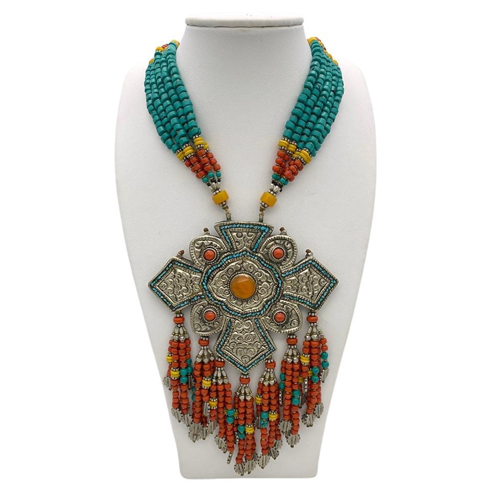 This is a statement necklace with Nepal plaque and fringes. This ethnic style colorful silk strung beaded necklace focuses on a 3.5 x 4.5 inch traditional patterned silver Nepal plaque with turquoise inlay, bezal set coral and an amber center. It is