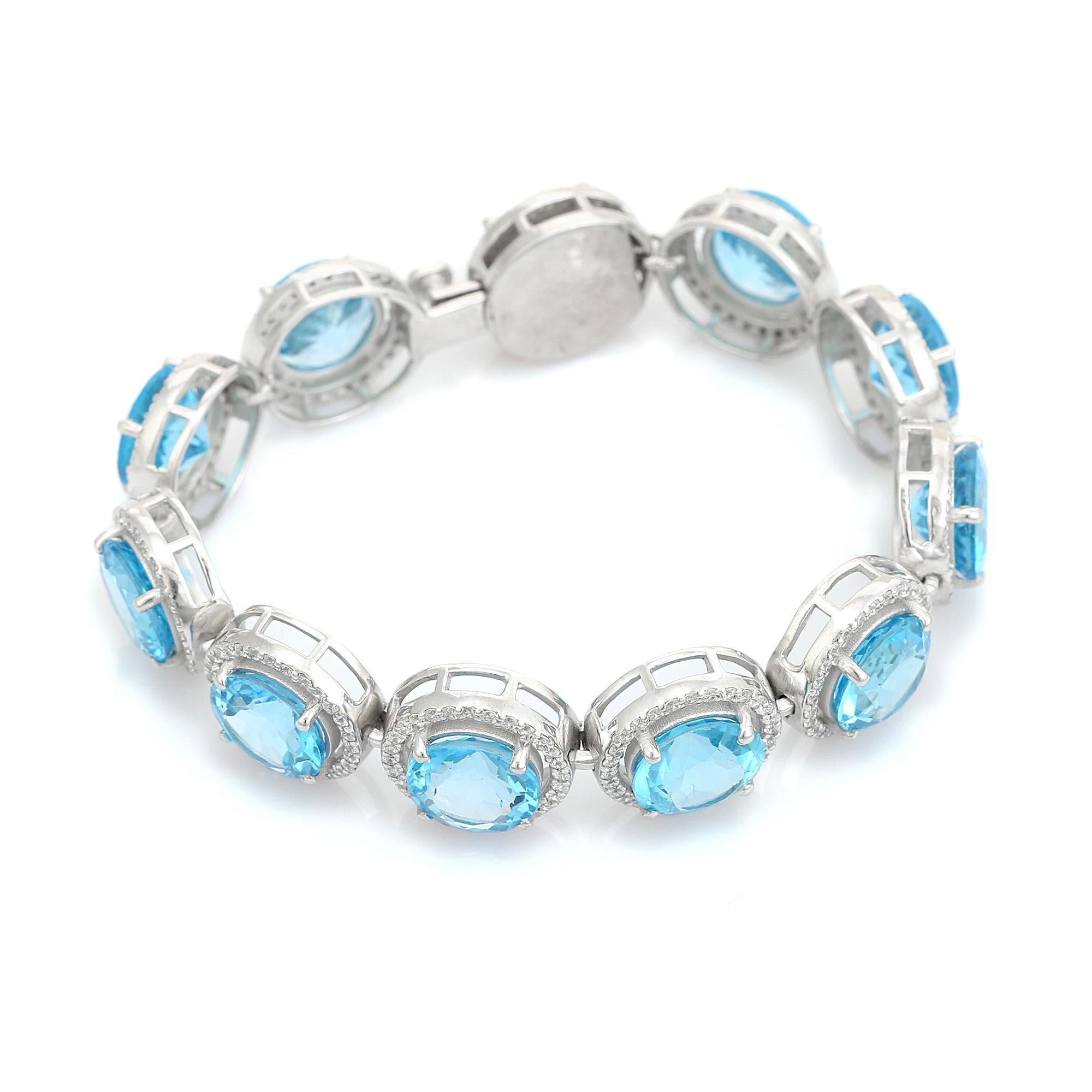 Esta impresionante pulsera de plata para bodas de topacio azul redondo con halo de circonitas presenta una hilera sin fisuras de gemas de topacio azul de talla redonda, cada una rodeada por un delicado halo de piedras blancas de acento, todo ello