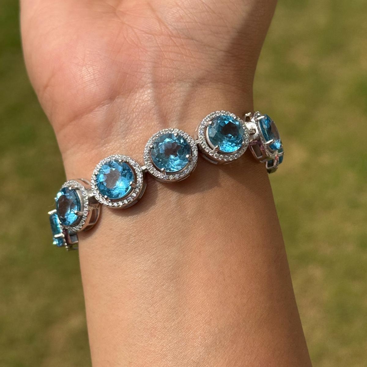 Questo splendido bracciale in argento con topazio blu rotondo e zirconi a raggiera presenta una fila ininterrotta di gemme di topazio blu a taglio rotondo, ciascuna circondata da una delicata aureola di pietre bianche, il tutto incastonato in un