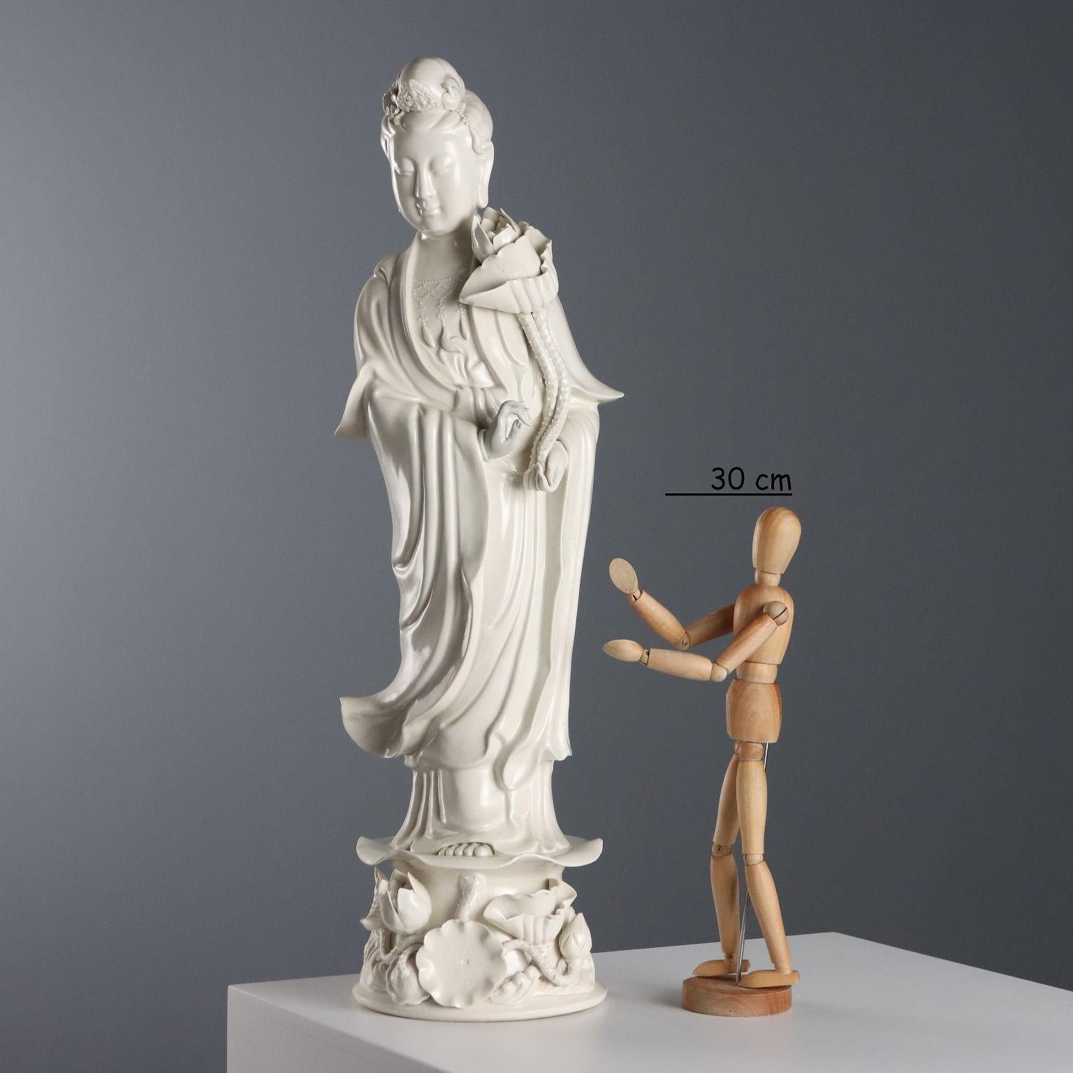 Scultura in blanc de chine raffigurante Guanyin con fiore di loto. La figura eretta poggia su una base a bordo mosso con decoro a rilievo di fiori e foglie. 