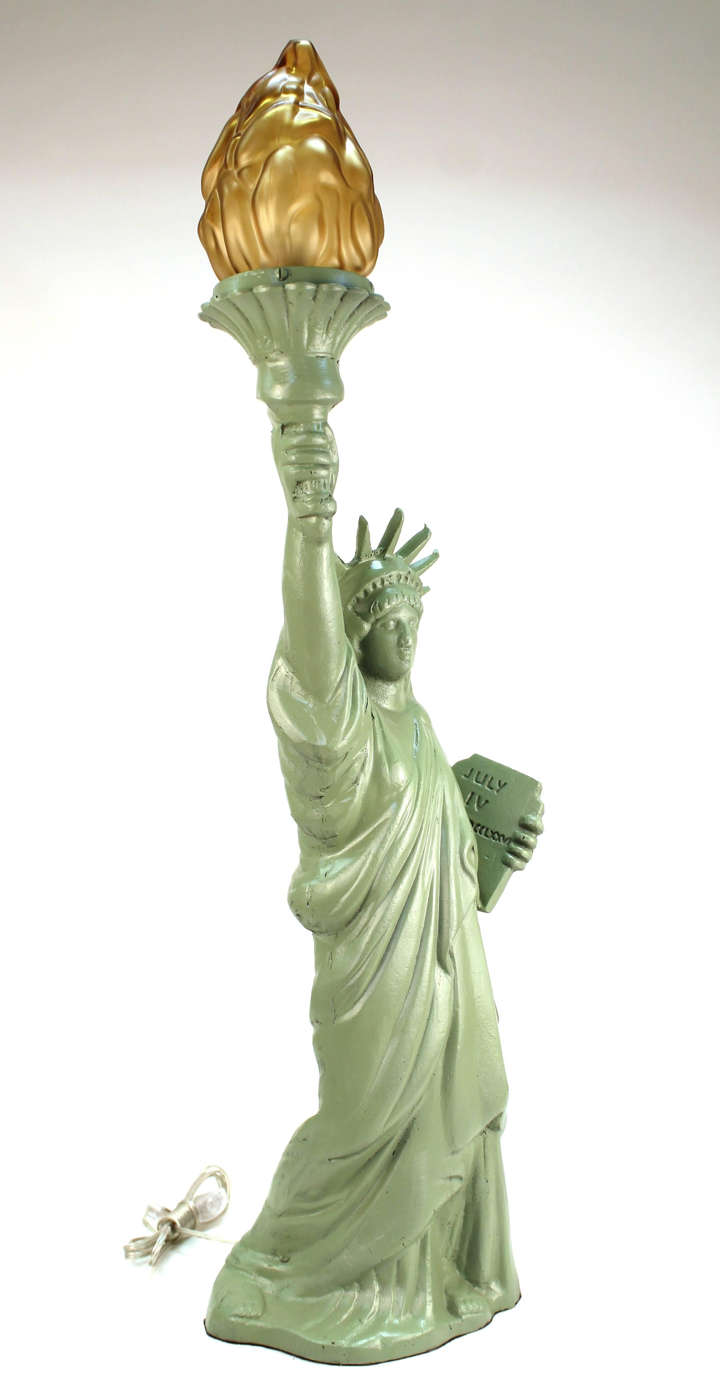 A table lamp in shape of the Statue of Liberty, made of green painted iron, with a glass flame in its torch. The base of the torch is marked 'Mexico'. The piece is in great vintage condition with age appropriate wear.