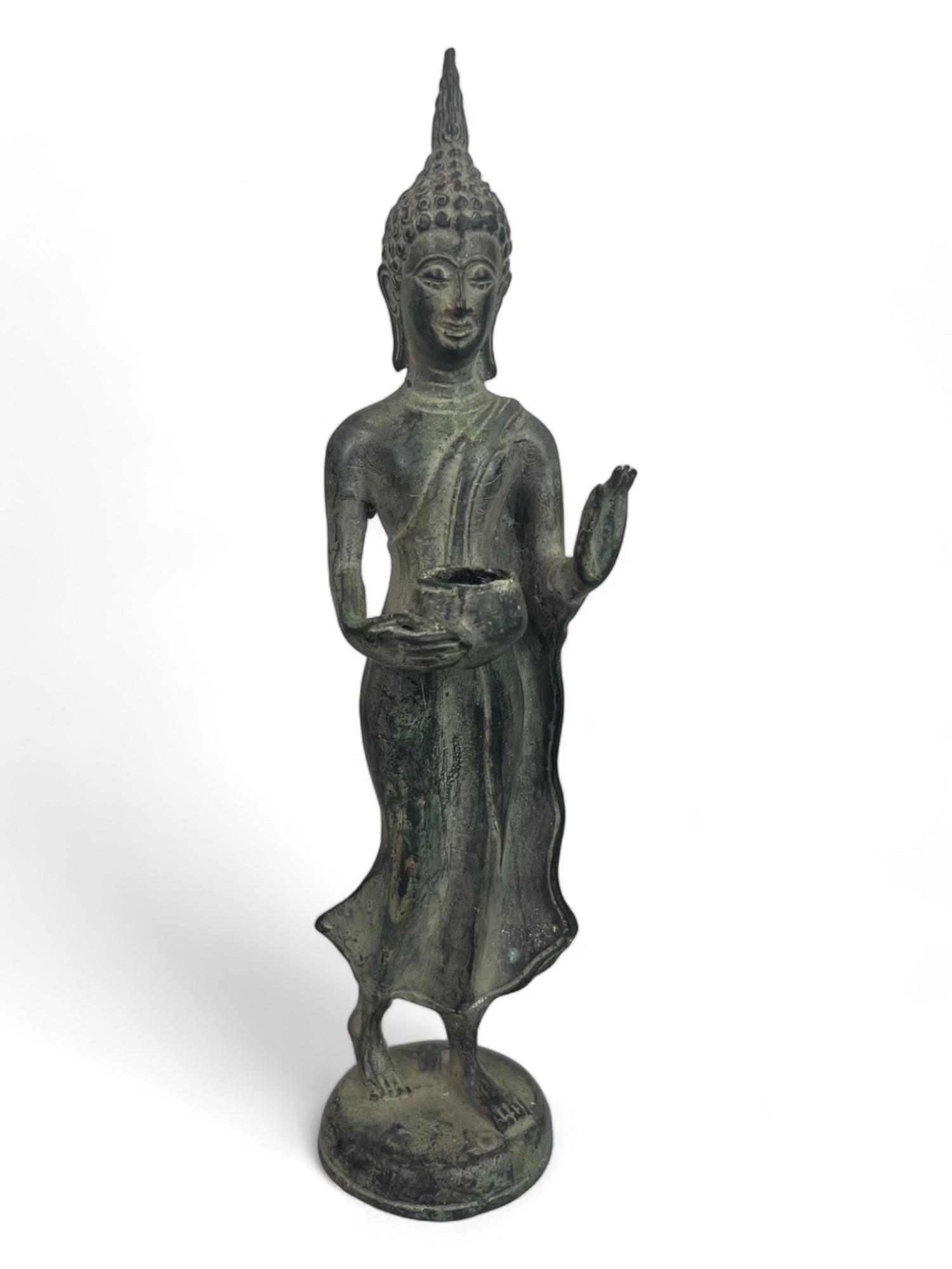 Statuette de Bouddha en bronze patiné, représenté en posture de marche, tenant un bol à aumônes dans la main droite et la main gauche levée, l'index et le pouce joints.
Cette position de la main est le geste de l'argumentation, de l'explication de