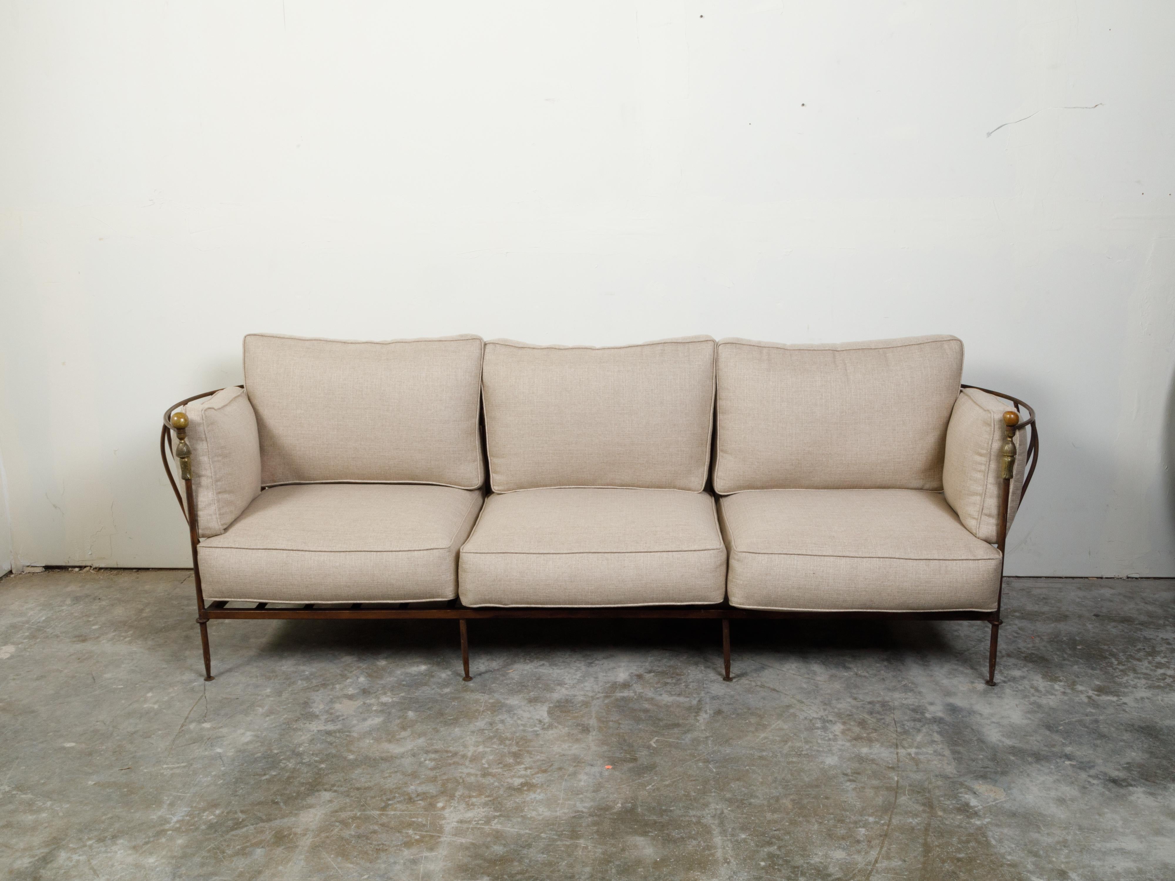 An iron and brass sofa from the mid 20th century possibly by Michael Taylor, with linen upholstery. Created during the midcentury period, this three-seat sofa features an iron structure with curving arm supports, accented with brass spheres and