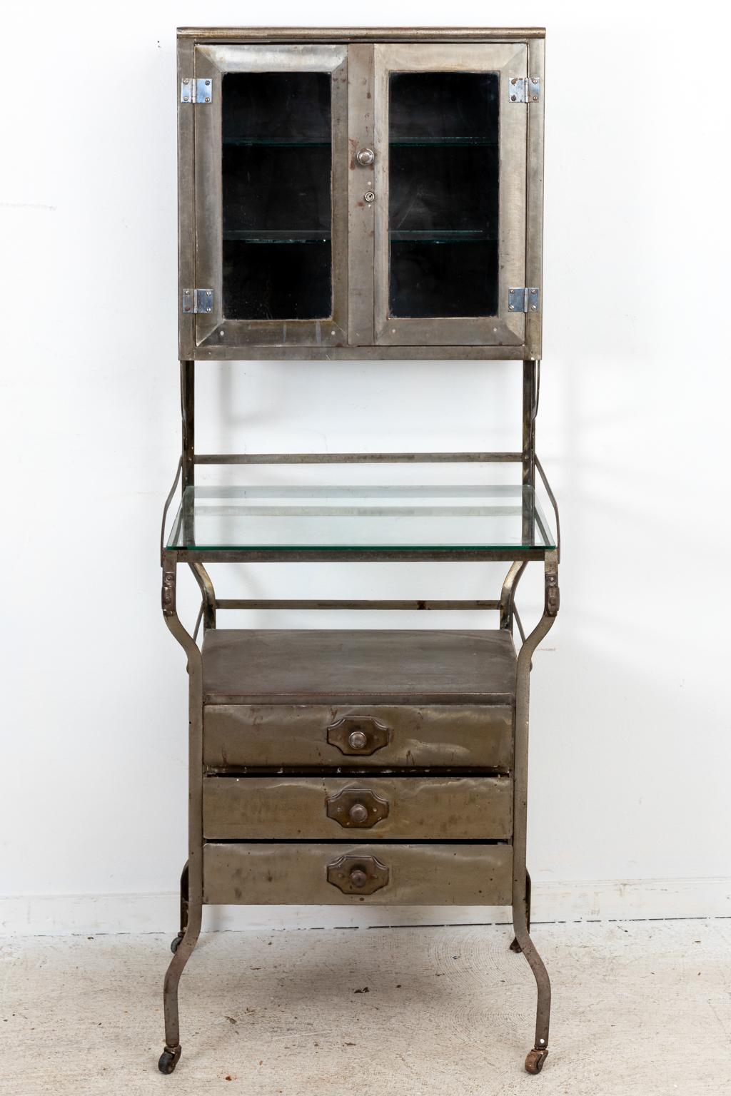 Industrial style steel and glass multi-level storage cabinet with round pulls. The piece is constructed with a glass front cabinet on the top, a central glass top work table, and a steel three drawer chest on the base for further storage. Between